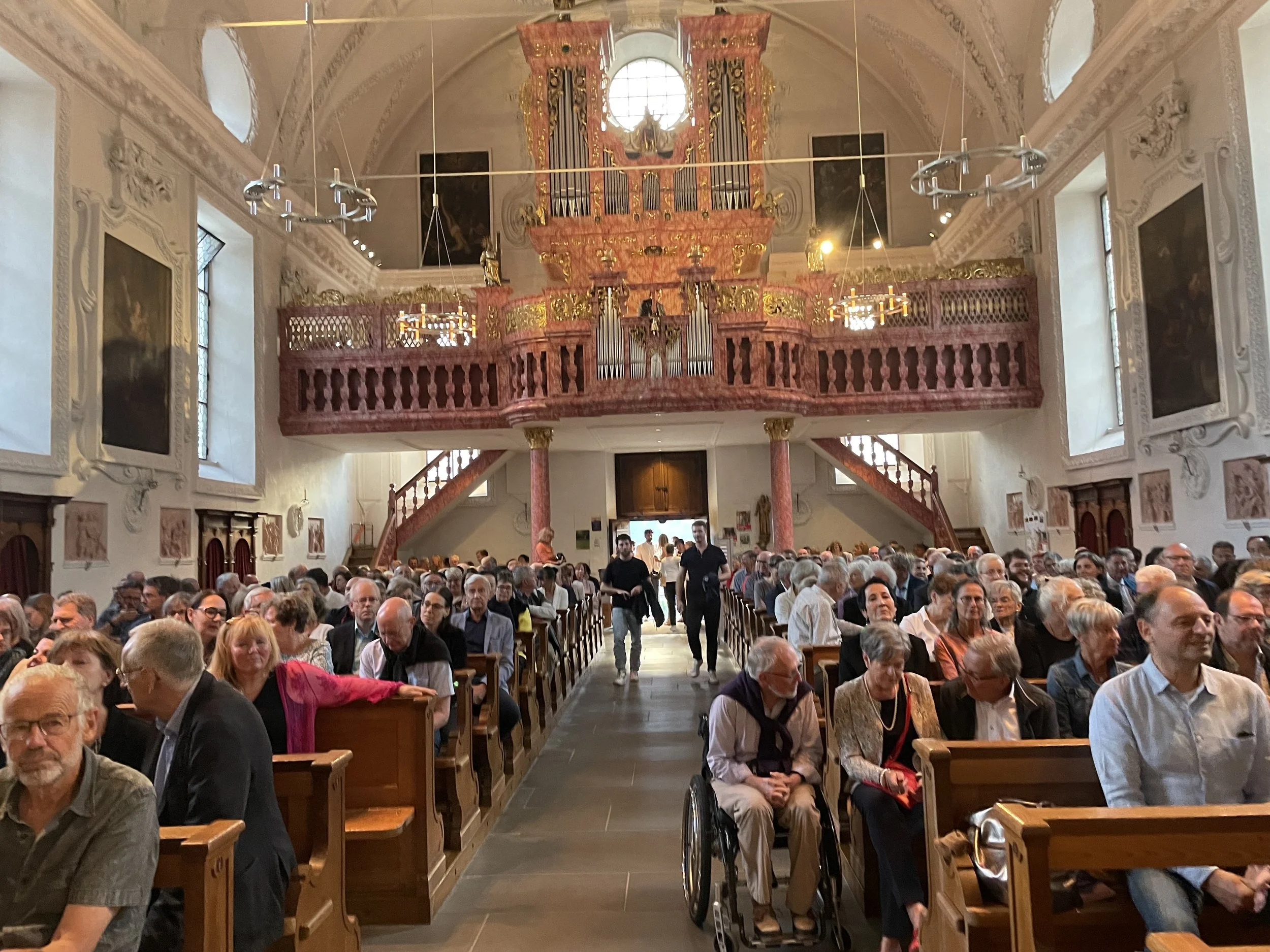 An interior of a church with a large congregation seated on wooden pews. There are two staircases leading up to an upper balcony, which is decorated with pink and gold accents. A large pipe organ is visible above the balcony, and the walls are adorne