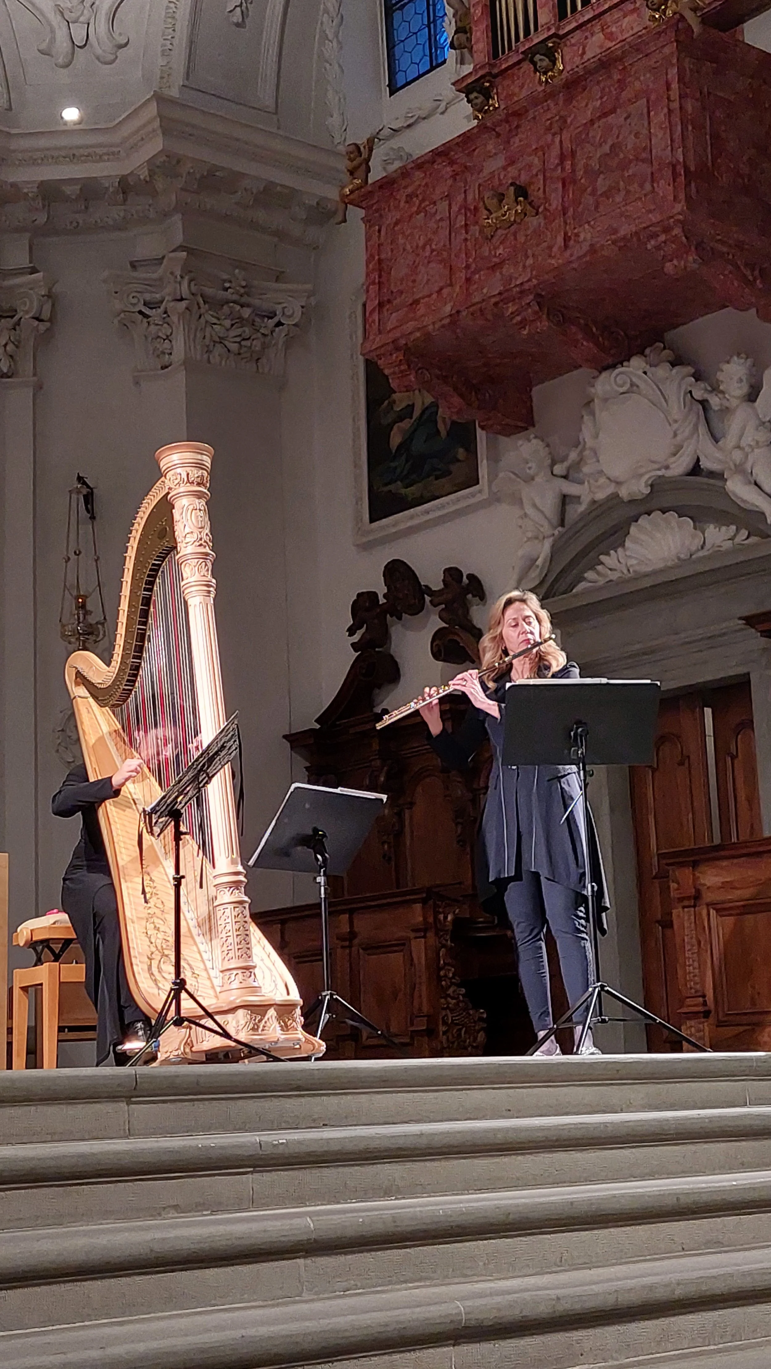 Two musicians perform on a stage in a historic concert hall, one playing a harp and the other a flute, with ornate architectural details behind them.