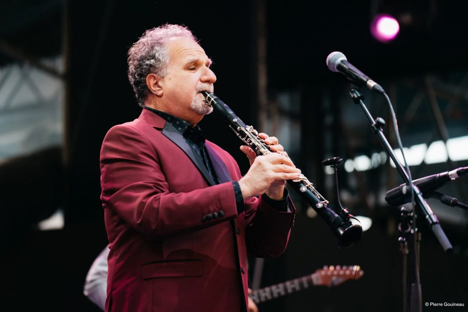 Man playing a clarinet on stage, wearing a maroon blazer and black shirt, with microphone in front of him.