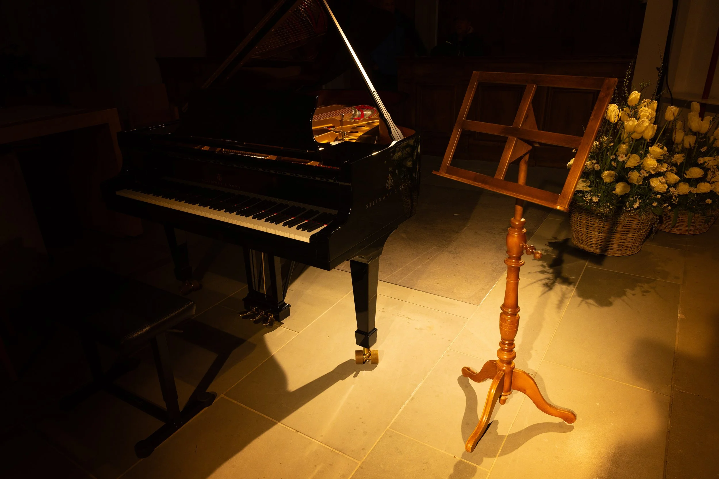 A grand piano, an empty music stand, and a basket of yellow flowers in a dimly lit room.