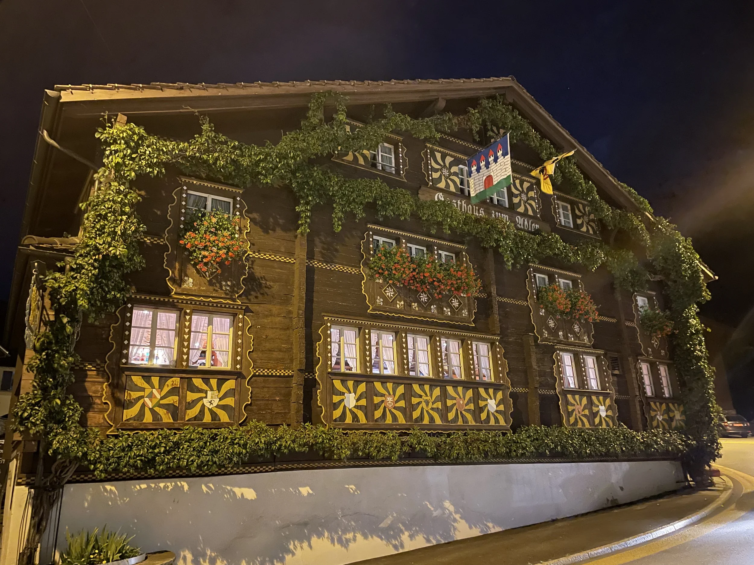 Nighttime view of a traditional Alpine house decorated with flower boxes, greenery, and a painted shield on the facade.
