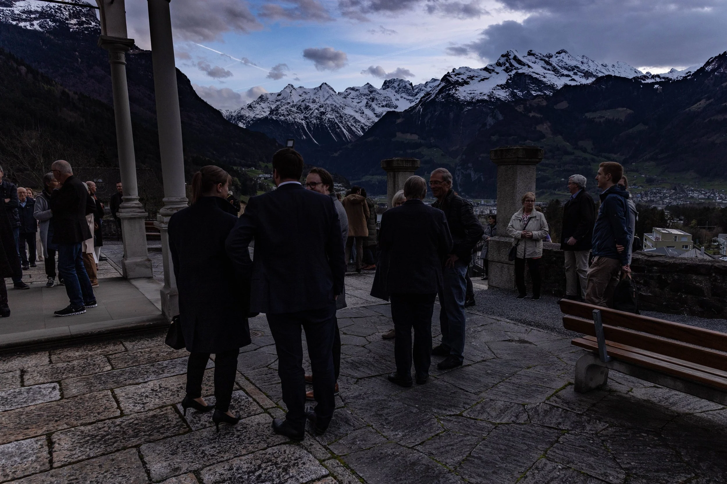People gathered outdoors on a terrace with mountains and snowy peaks in the background during dusk or evening.