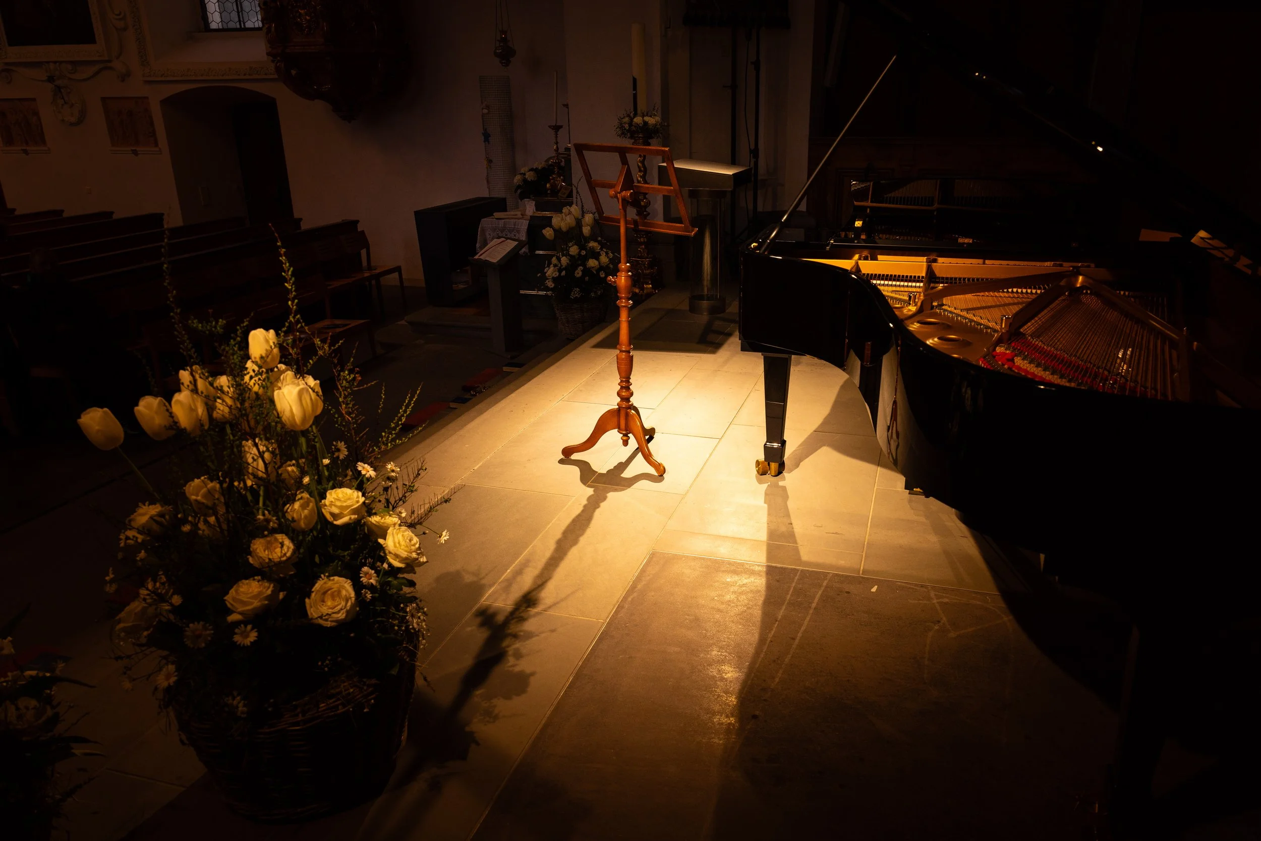 A grand piano with its lid open, a wooden music stand, a floral arrangement, and a candle on the altar in a dimly lit church or concert hall.