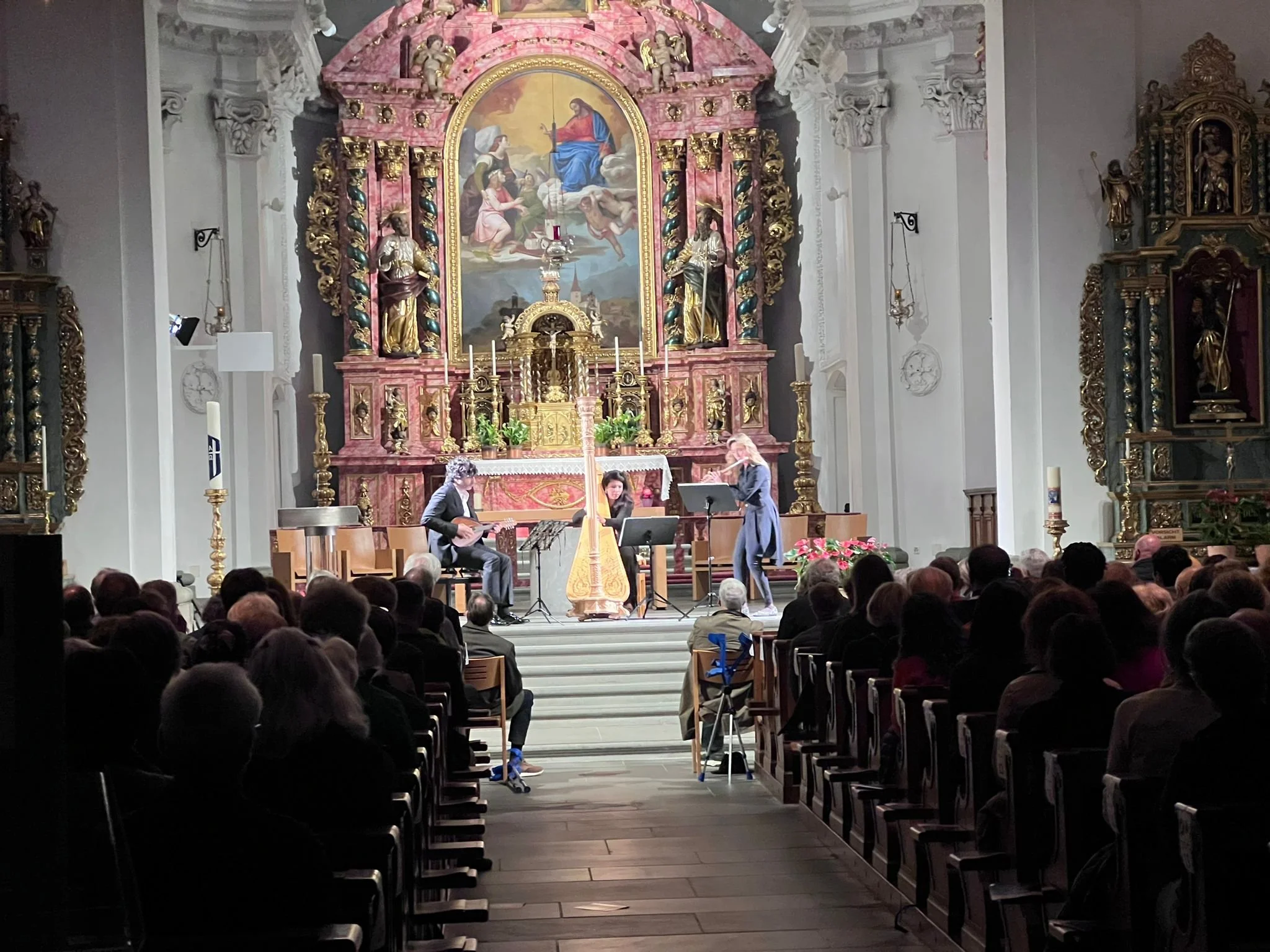 A classical music performance is taking place inside a church with ornate gold and pink altar in the background, and an audience seated facing the stage.