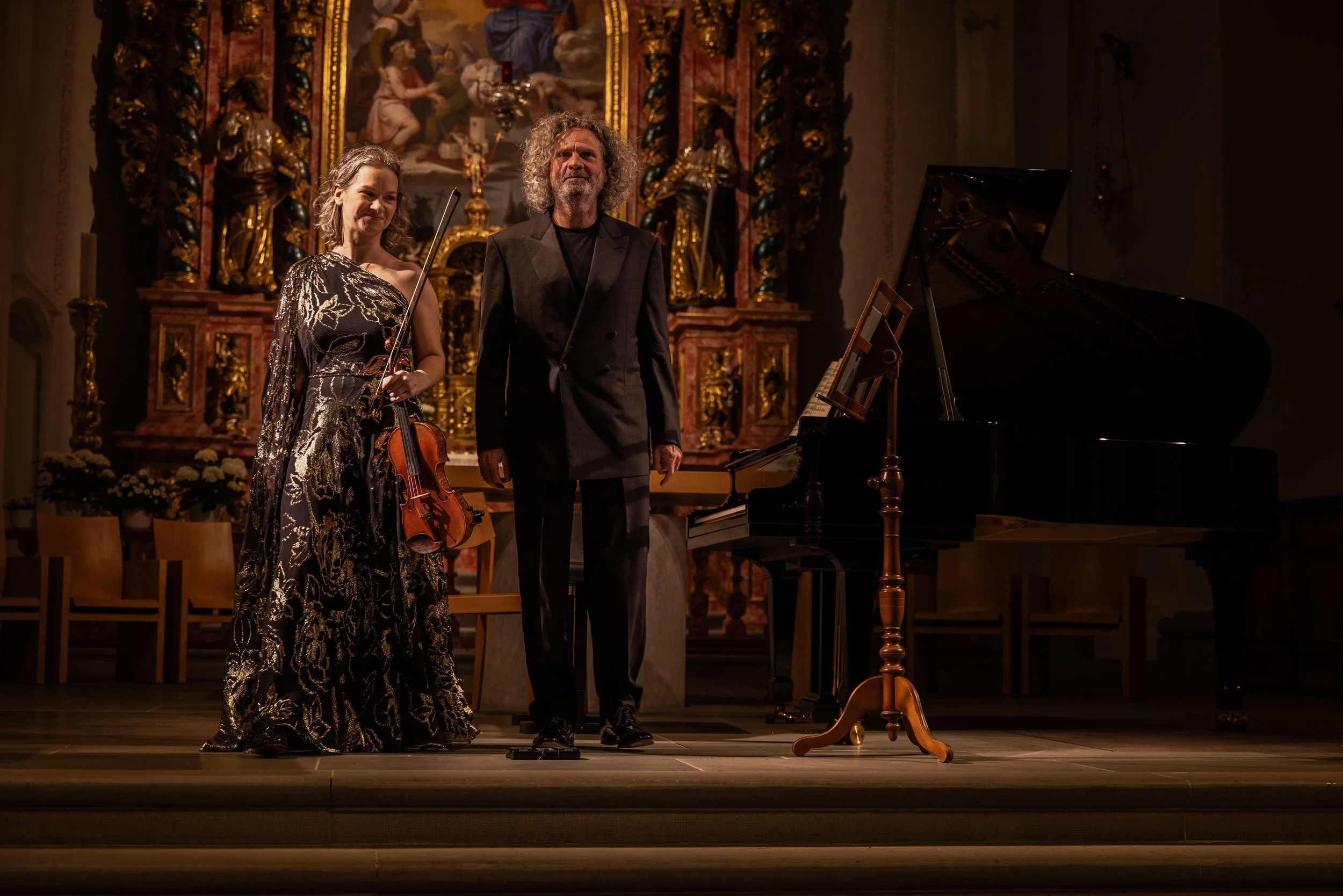 Two musicians, a woman with a violin and a man with a bow, standing on stage after a performance, with a grand piano beside them and a decorated religious altar in the background.