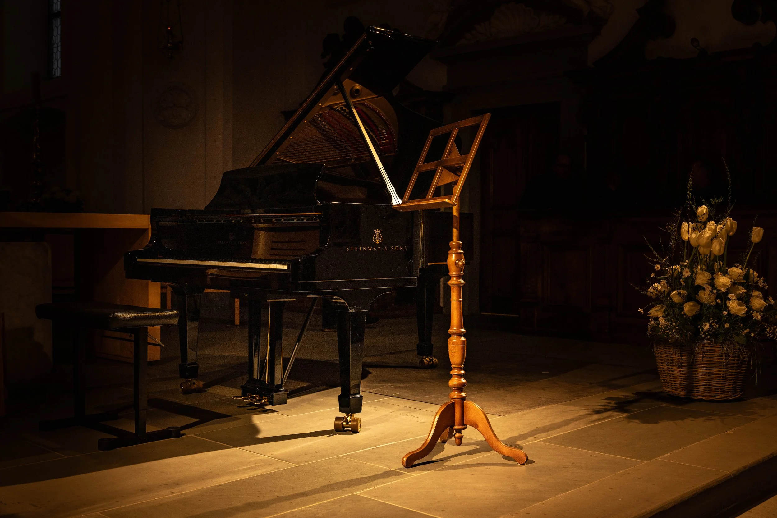 A grand piano with an open lid, a music stand, and a stool on a stage with a flower basket to the right.