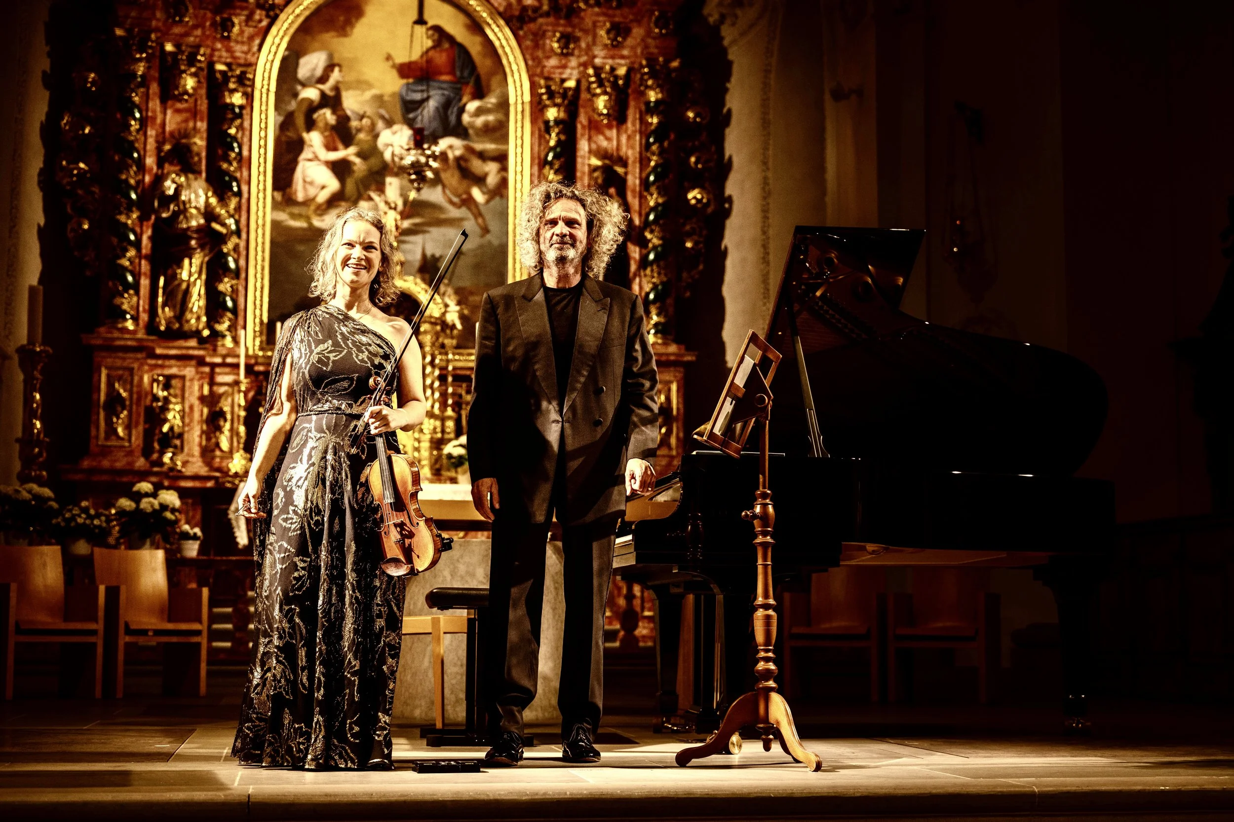 Two musicians, a woman holding a violin and a man standing beside her, on a stage in a church or concert hall, with an ornate religious altar and painting in the background.