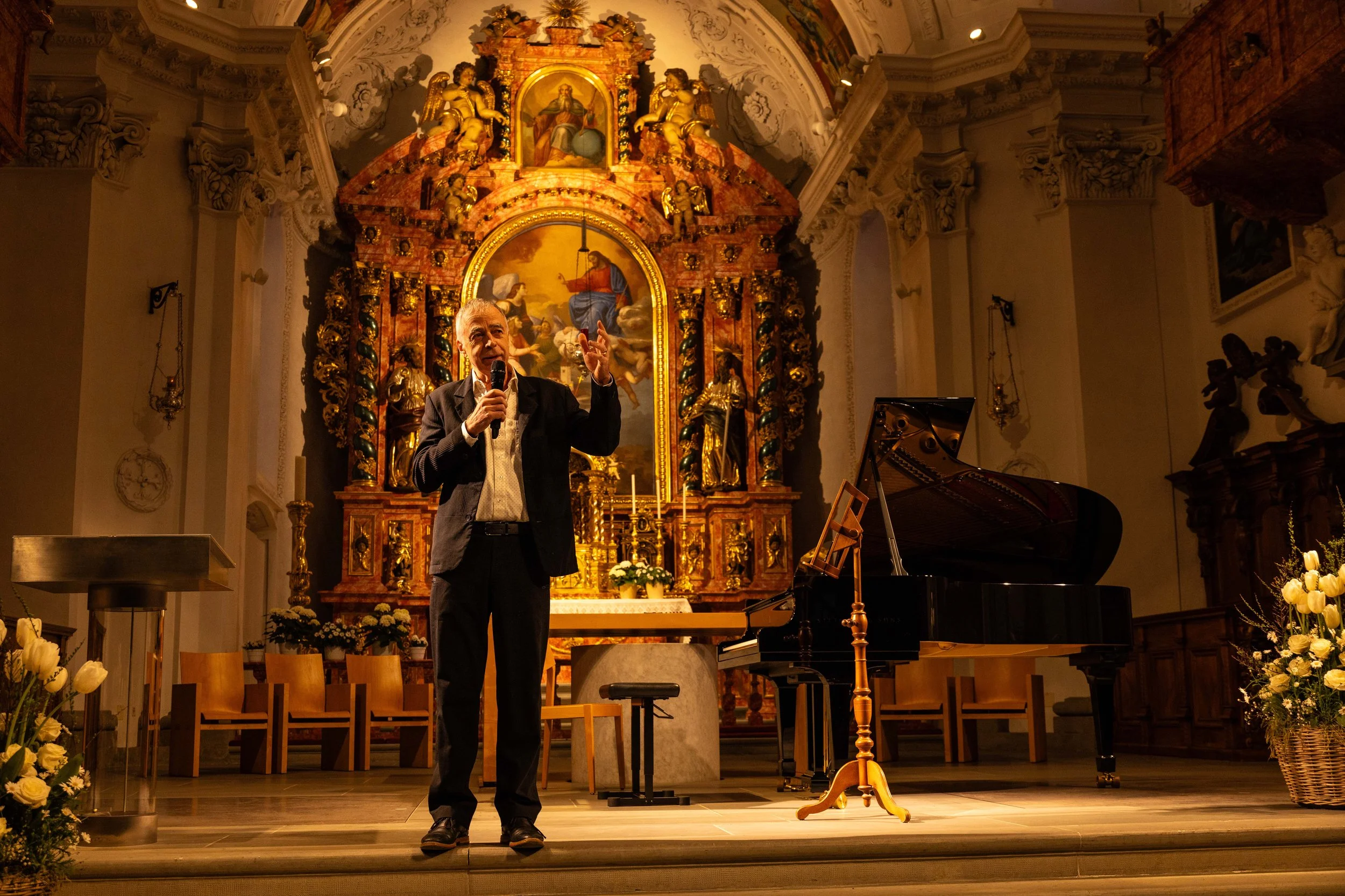 An older man speaking into a microphone on a stage in a church, with a grand altar, a piano, and floral arrangements in the background.