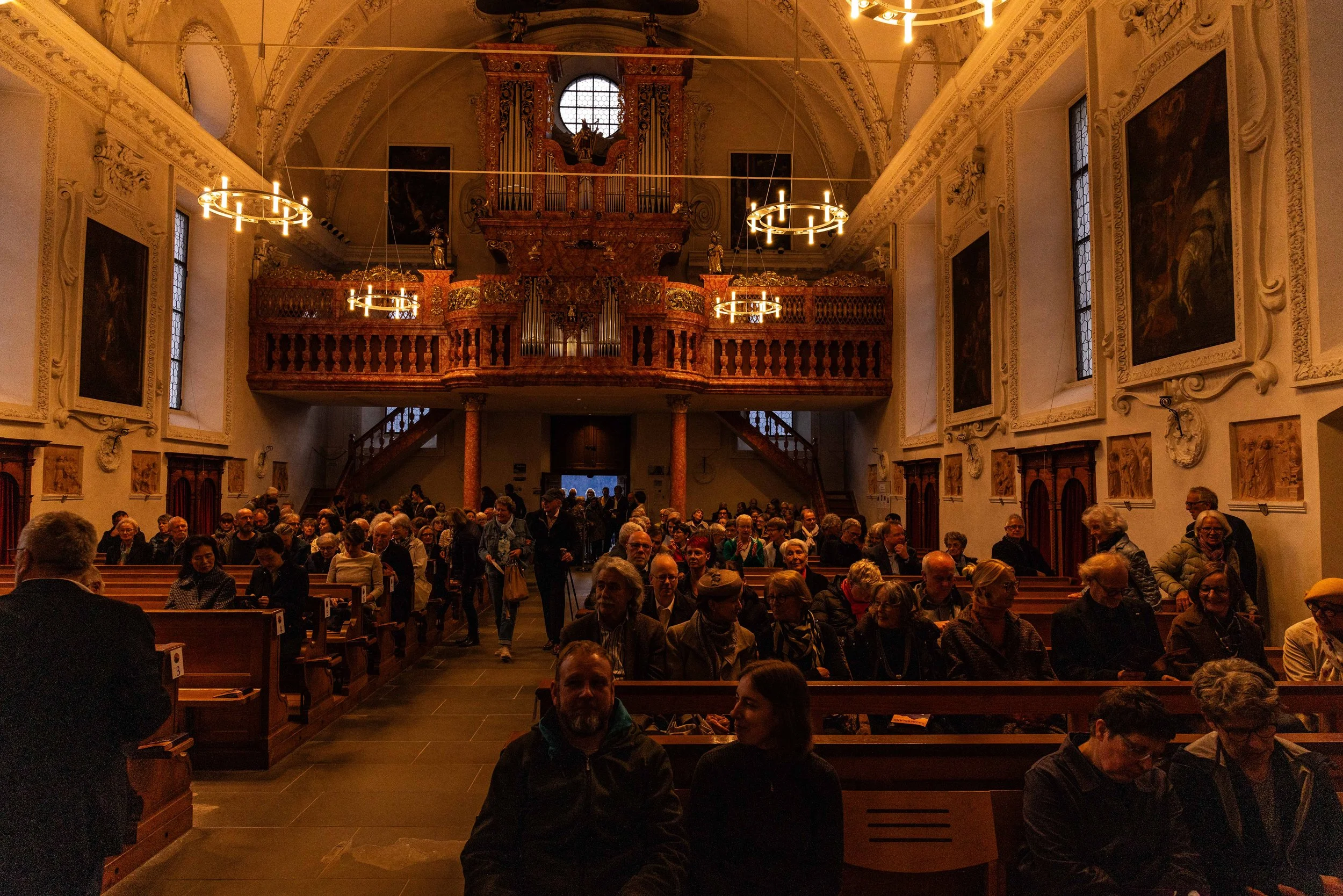 An interior view of a church or cathedral with many people seated in wooden pews, ornate wall decorations, paintings, and a large pipe organ at the back.