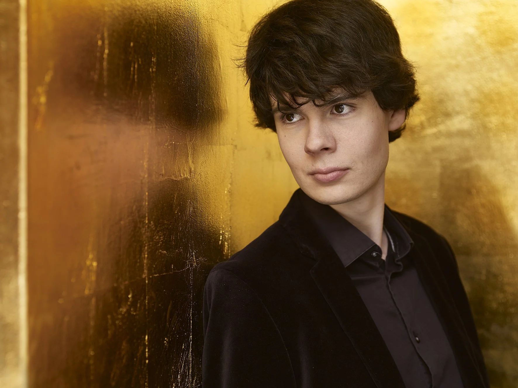 A young man with dark, wavy hair and light skin looking to his right, standing beside a shiny, gold-colored wall.