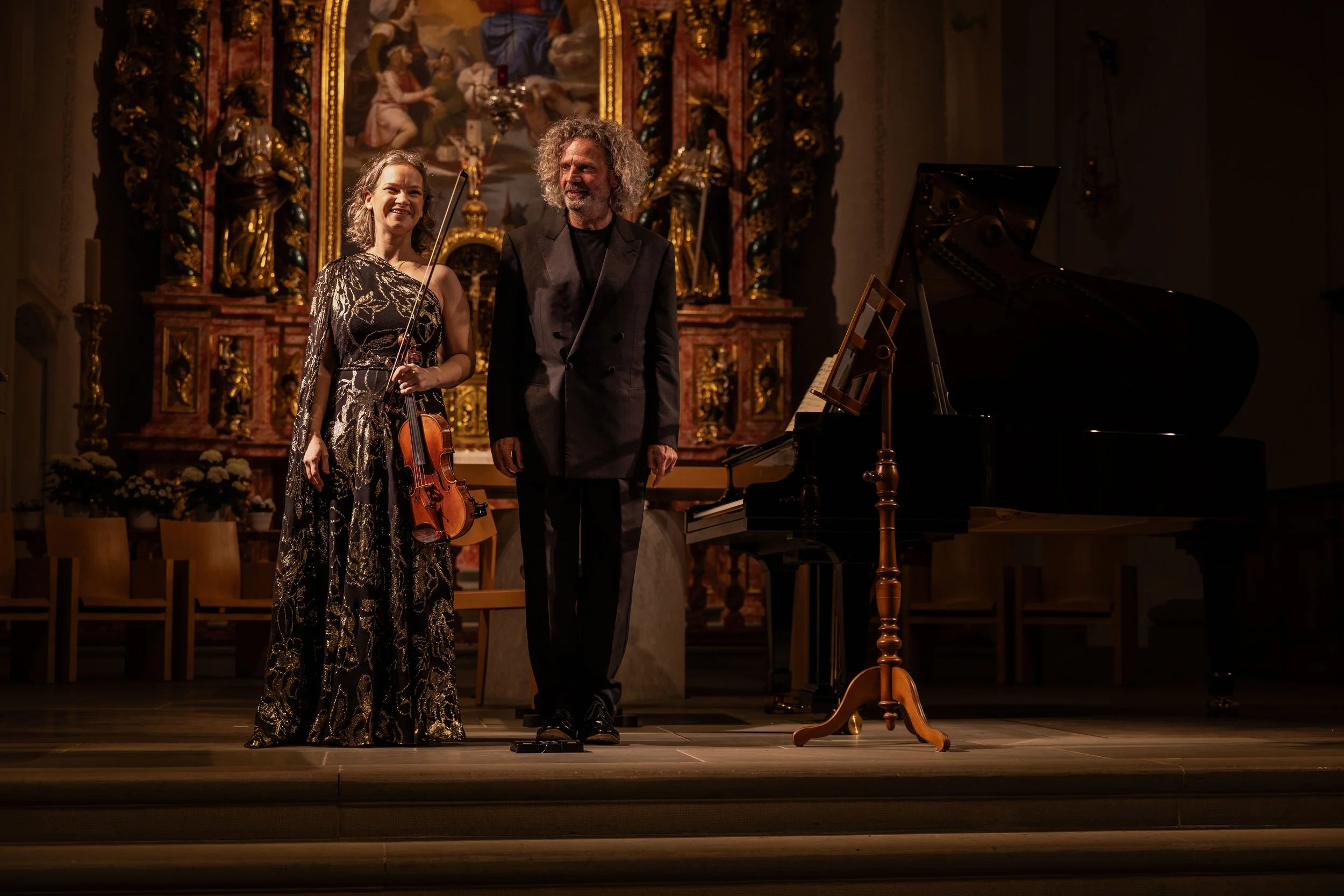 Two musicians, a woman holding a violin and a man, standing on a stage in a concert hall with elaborate gold and red decorations, a large religious painting in the background, and a black grand piano to the right.