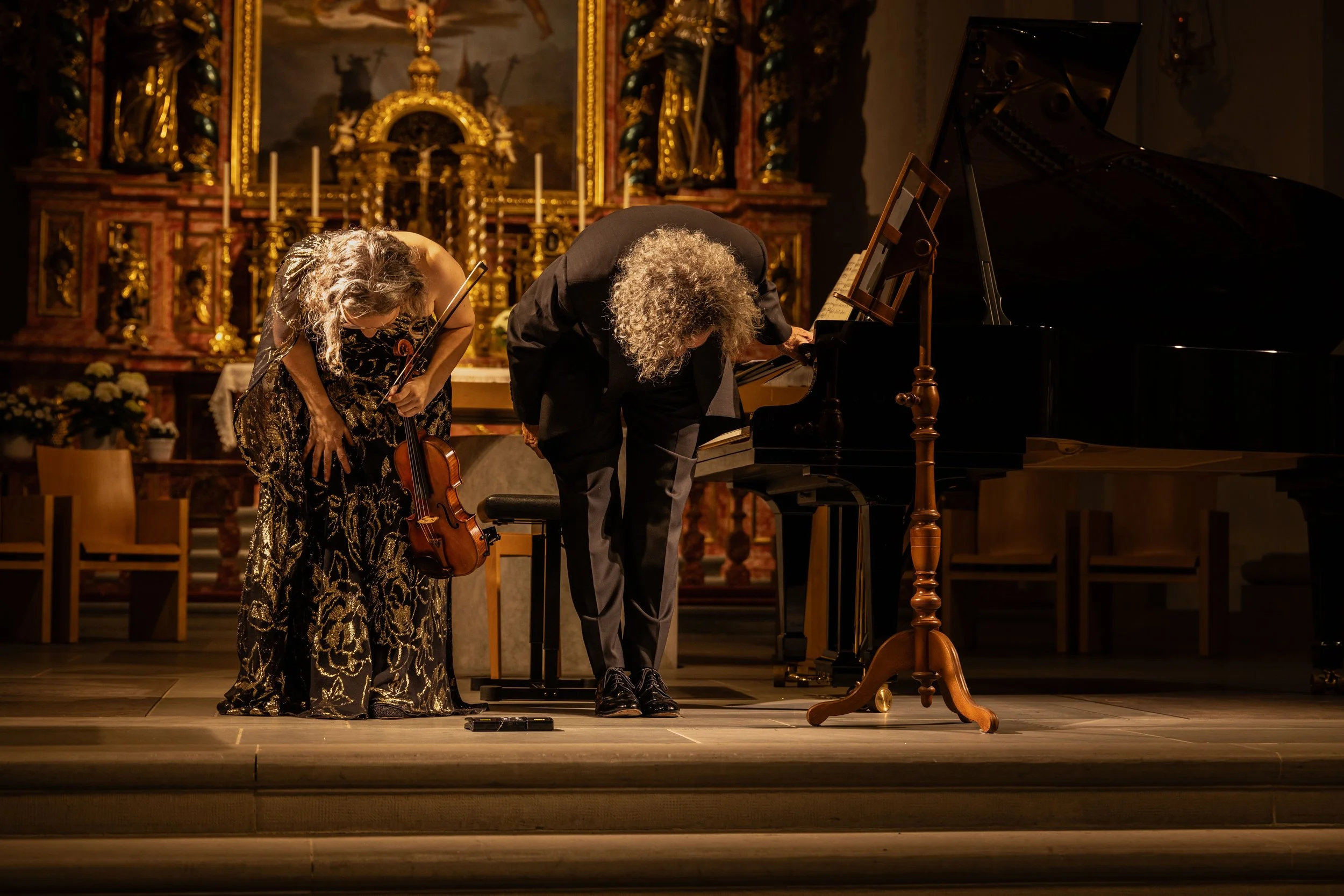Two musicians bowing after a performance on stage in a church setting, one holding a violin and the other a grand piano, with ornate religious decorations and candles in the background.