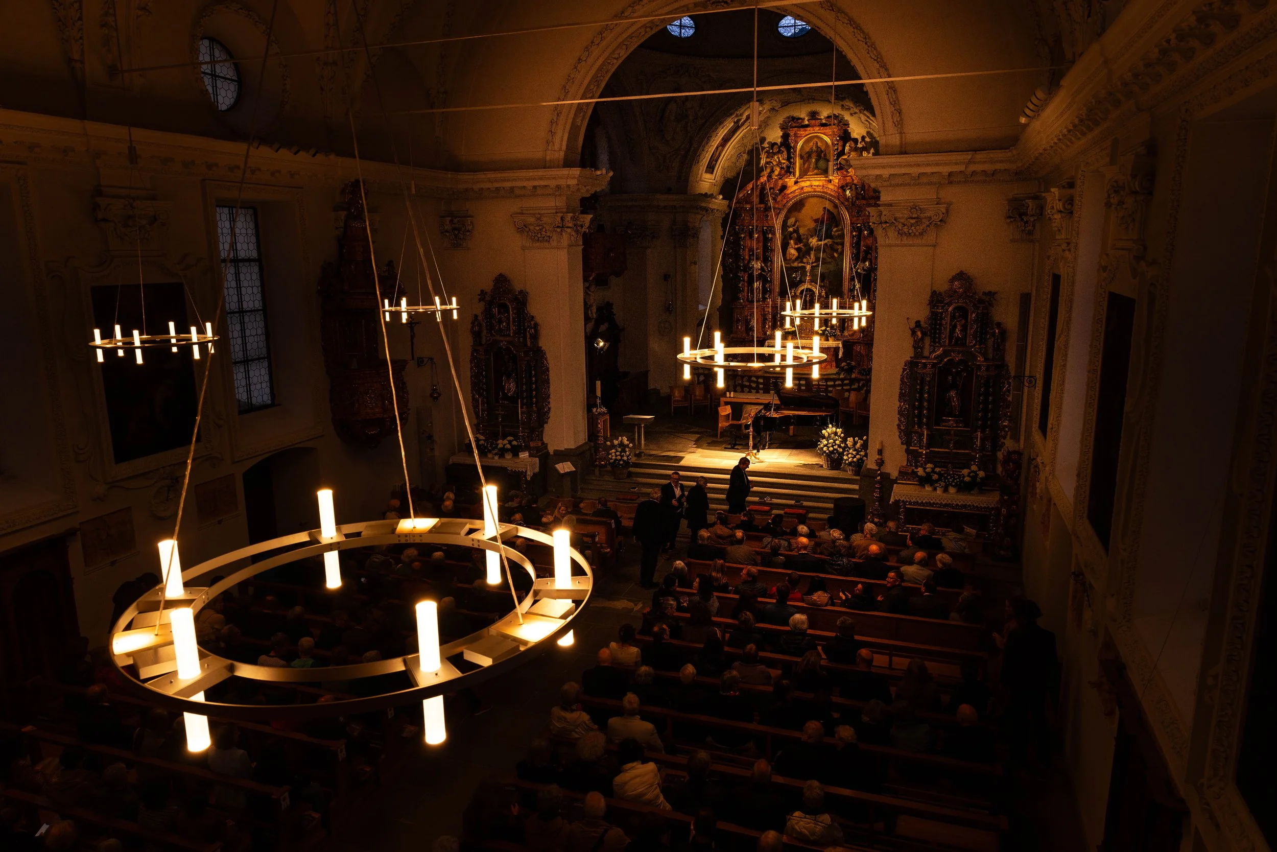 Inside a church with ornate decorations, a stage at the front, and many seated attendees. There are large circular chandeliers hanging from the ceiling.