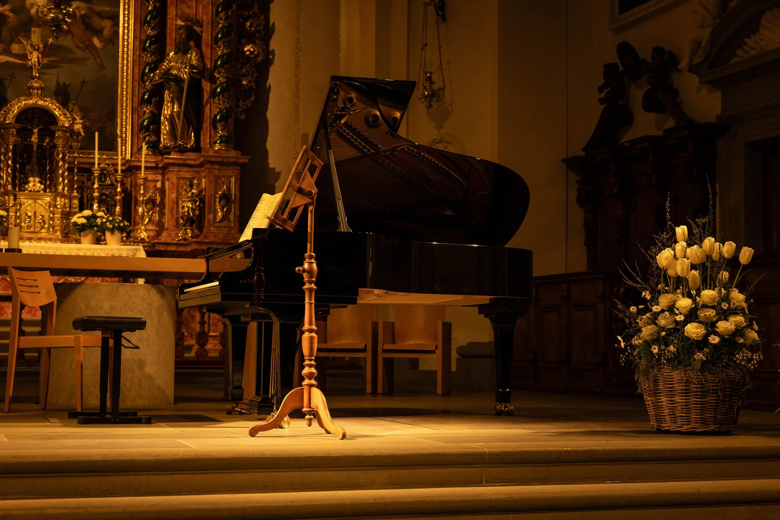A grand piano on a stage in a church or cathedral, with a wooden music stand, a flower arrangement in a wicker basket, and ornate religious decorations and artwork in the background.