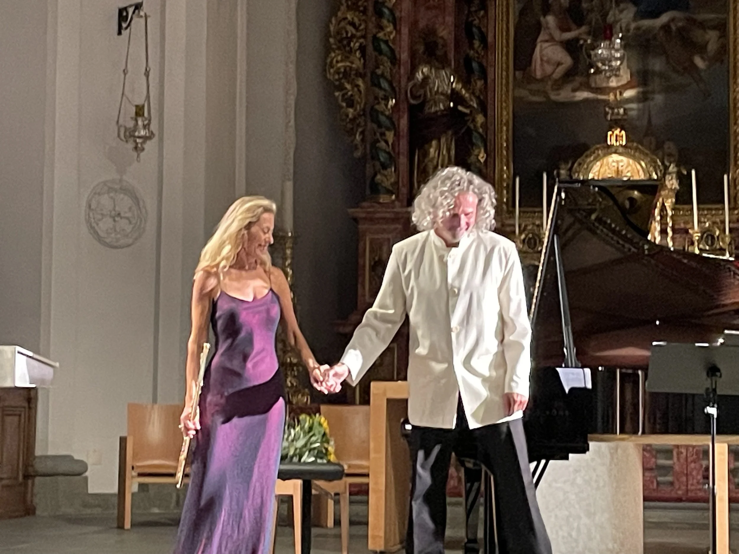 A woman in a purple dress holding a baton and a man with curly gray hair in a white coat holding her hand, standing on a stage with a grand piano and a religious setting in the background.
