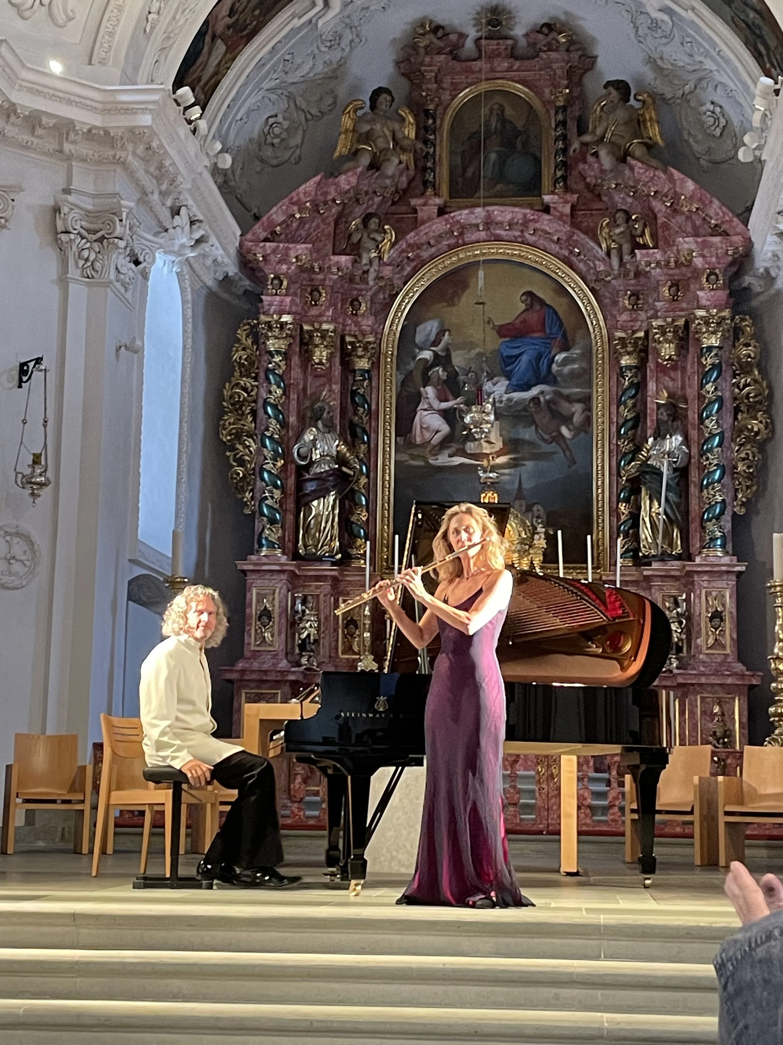 Two musicians performing in a church: a pianist seated at a grand piano and a woman standing and playing the flute, with ornate religious artwork and statues in the background.