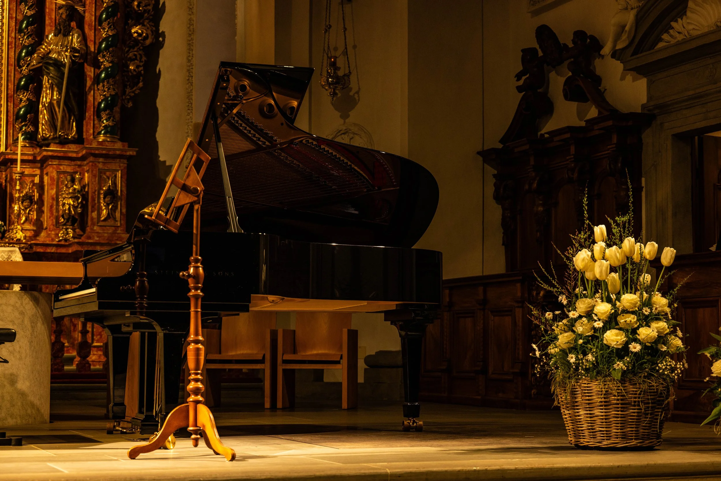 A grand piano on a wooden stage with a flower arrangement in a wicker basket to the right, and ornate church altar with religious icons in the background.
