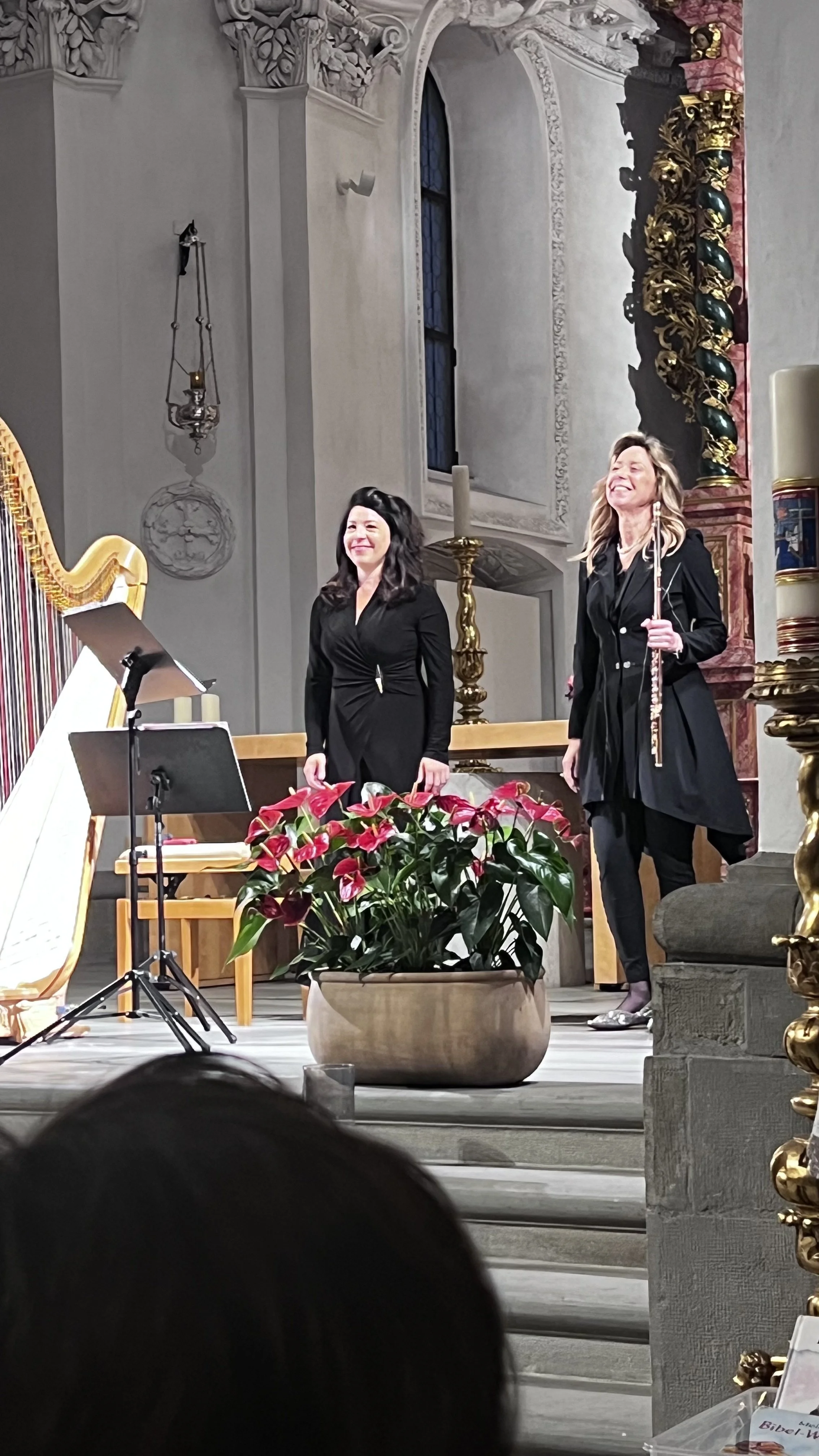 Two women standing on a church altar stage, one holding a flute, both smiling, with a large pink flowers in a pot in front of them. The background features ornate church interior details with high windows and decorative elements.
