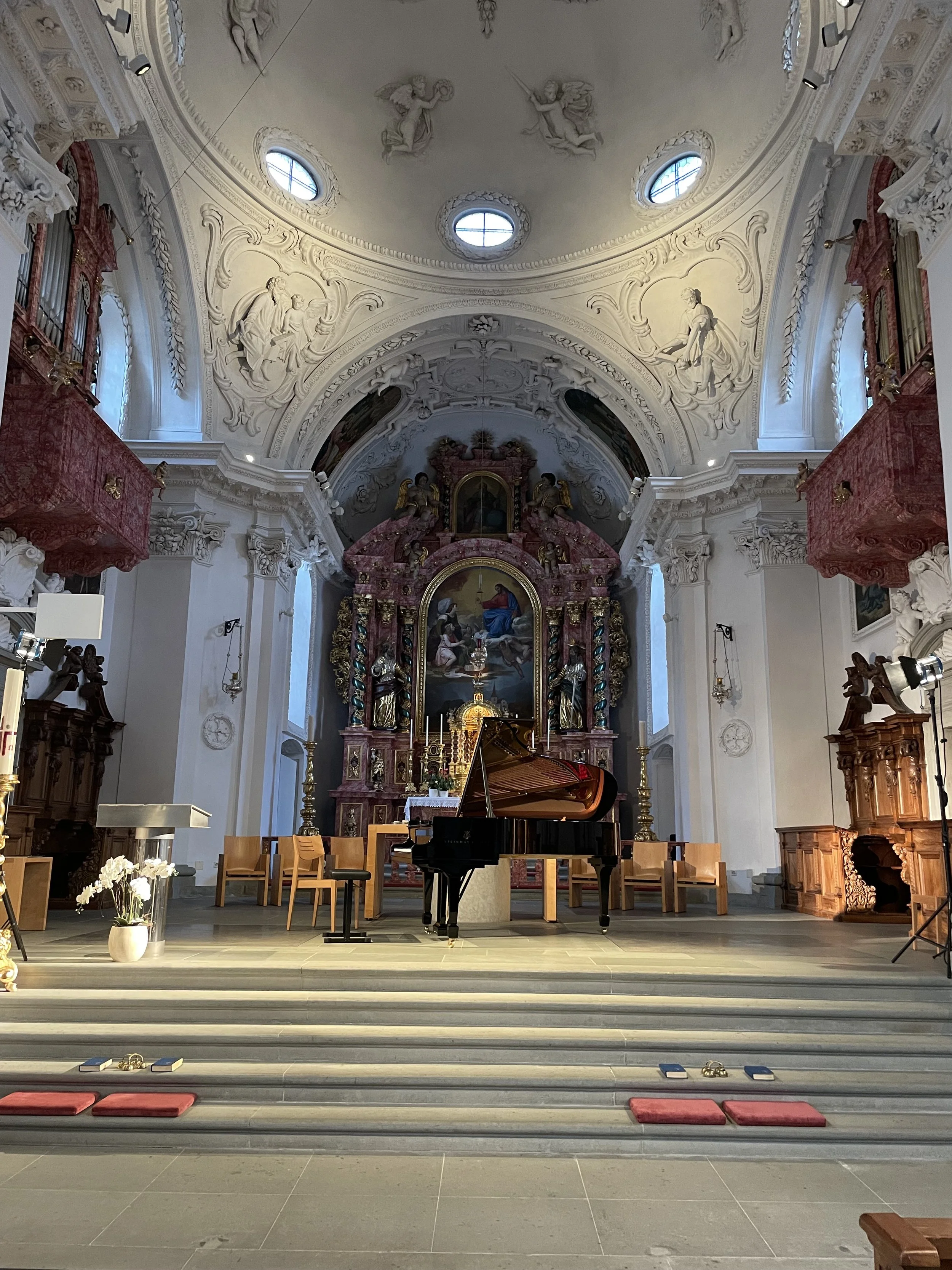 Interior of a church with a grand red and gold altar, a black grand piano in the center, and wooden chairs around it. The ceiling is ornate with white stucco sculptures and circular windows letting in natural light.