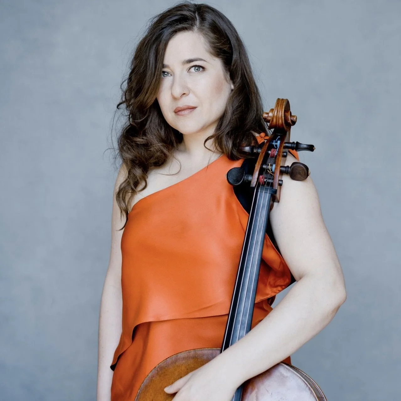 A woman with long brown curly hair, wearing an orange valued dress, holding a cello against a plain background.