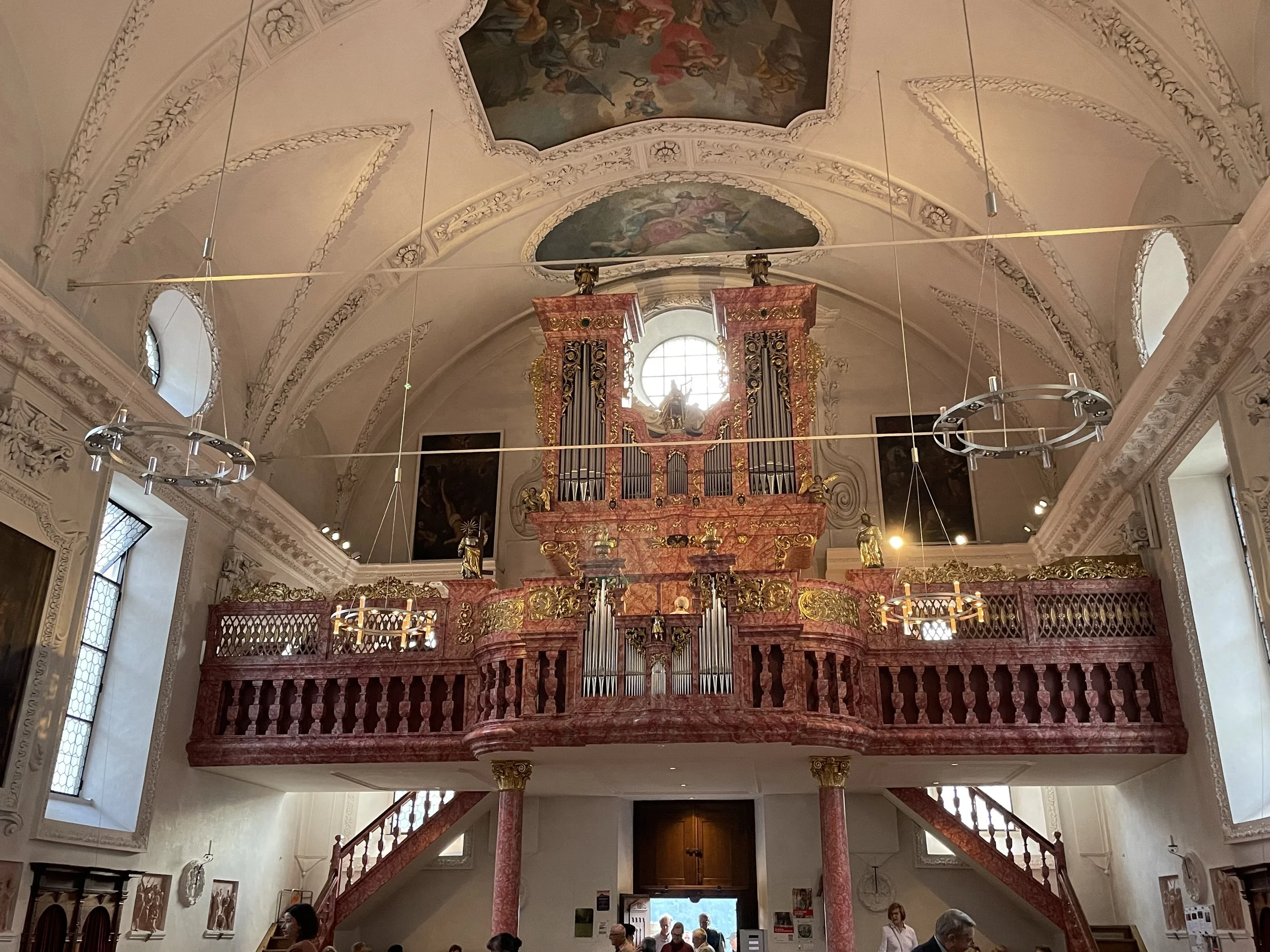 Interior of a church with ornate decorations, a large pipe organ on a balcony, and people standing inside.