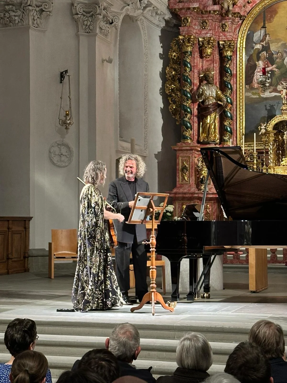 Two performers, a woman in a patterned black and gold gown and a man in a black suit, standing on a stage with a grand piano, in a decorated church or concert hall, engaging with the audience.