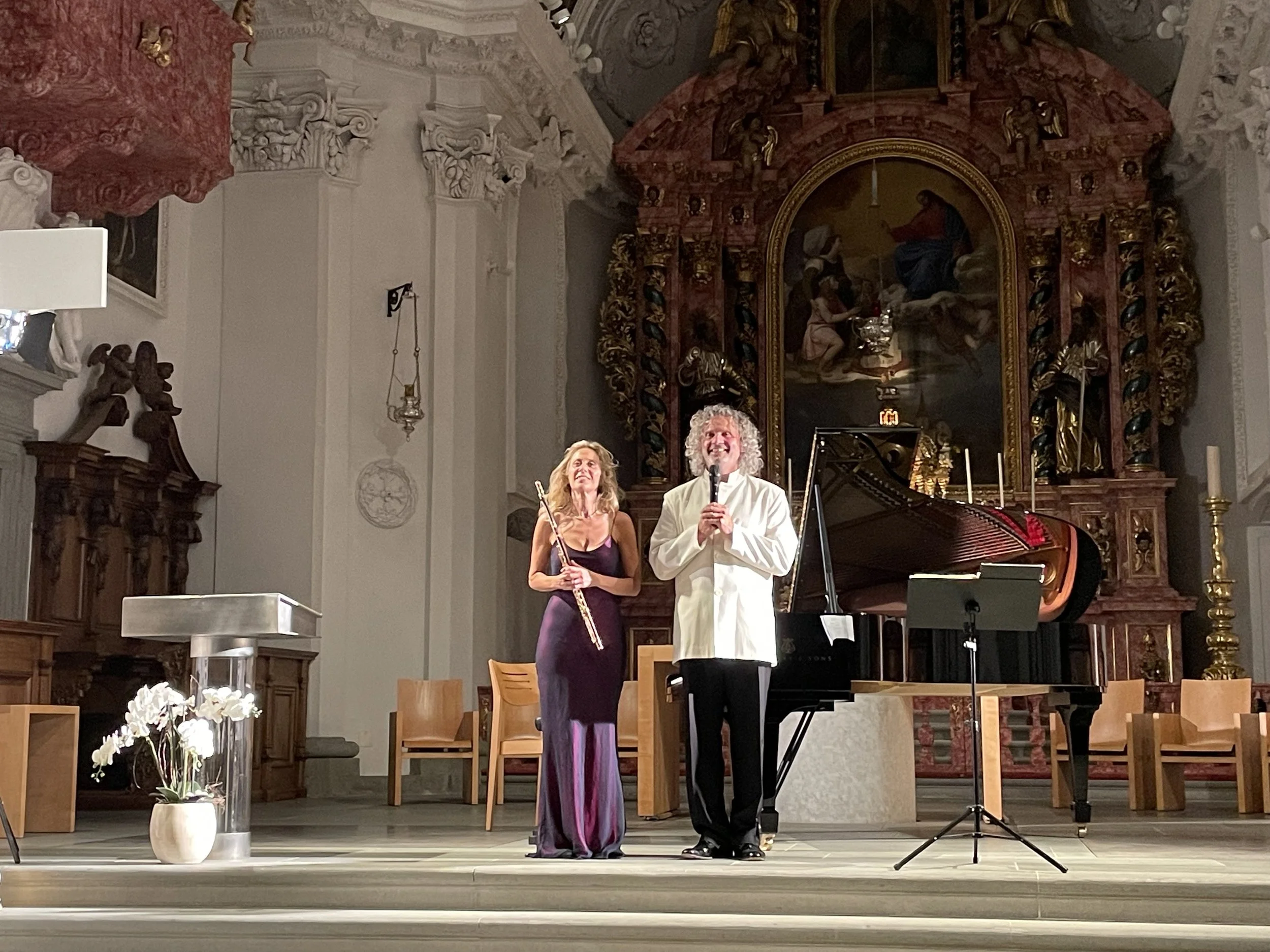 Two musicians perform on stage inside a church or concert hall, with a grand piano and a large, ornate religious altarpiece in the background. One woman holds a flute and wears a purple evening gown, while a man in a white jacket holds a microphone.
