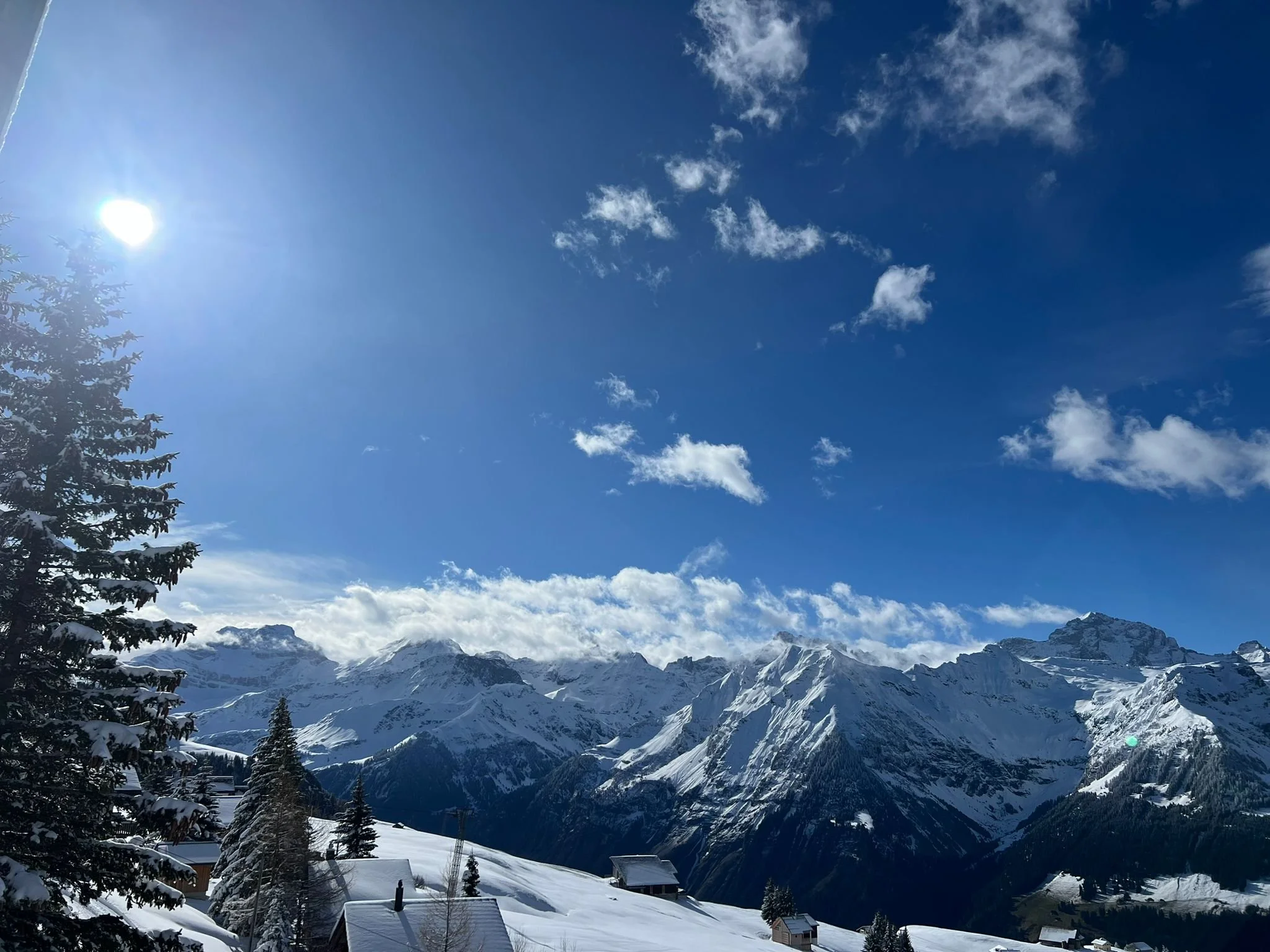 Snow-covered mountain range under a bright blue sky with scattered clouds and the sun shining.