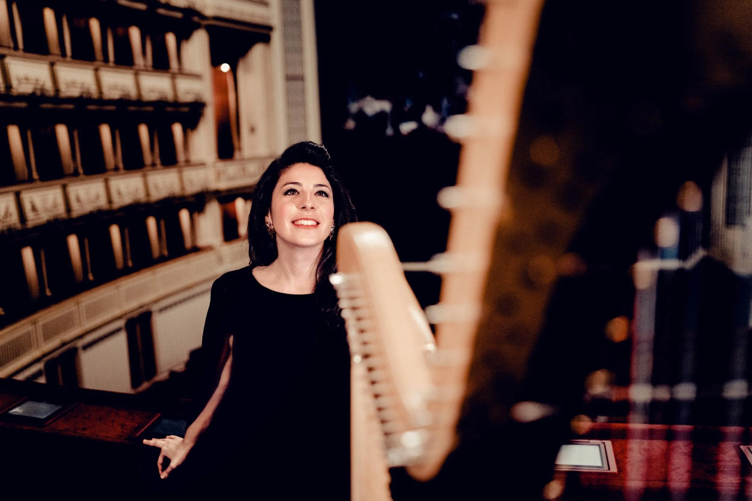 A woman with dark hair smiling and looking up, seated at a piano with wood grain and strings visible, in a dimly lit room.