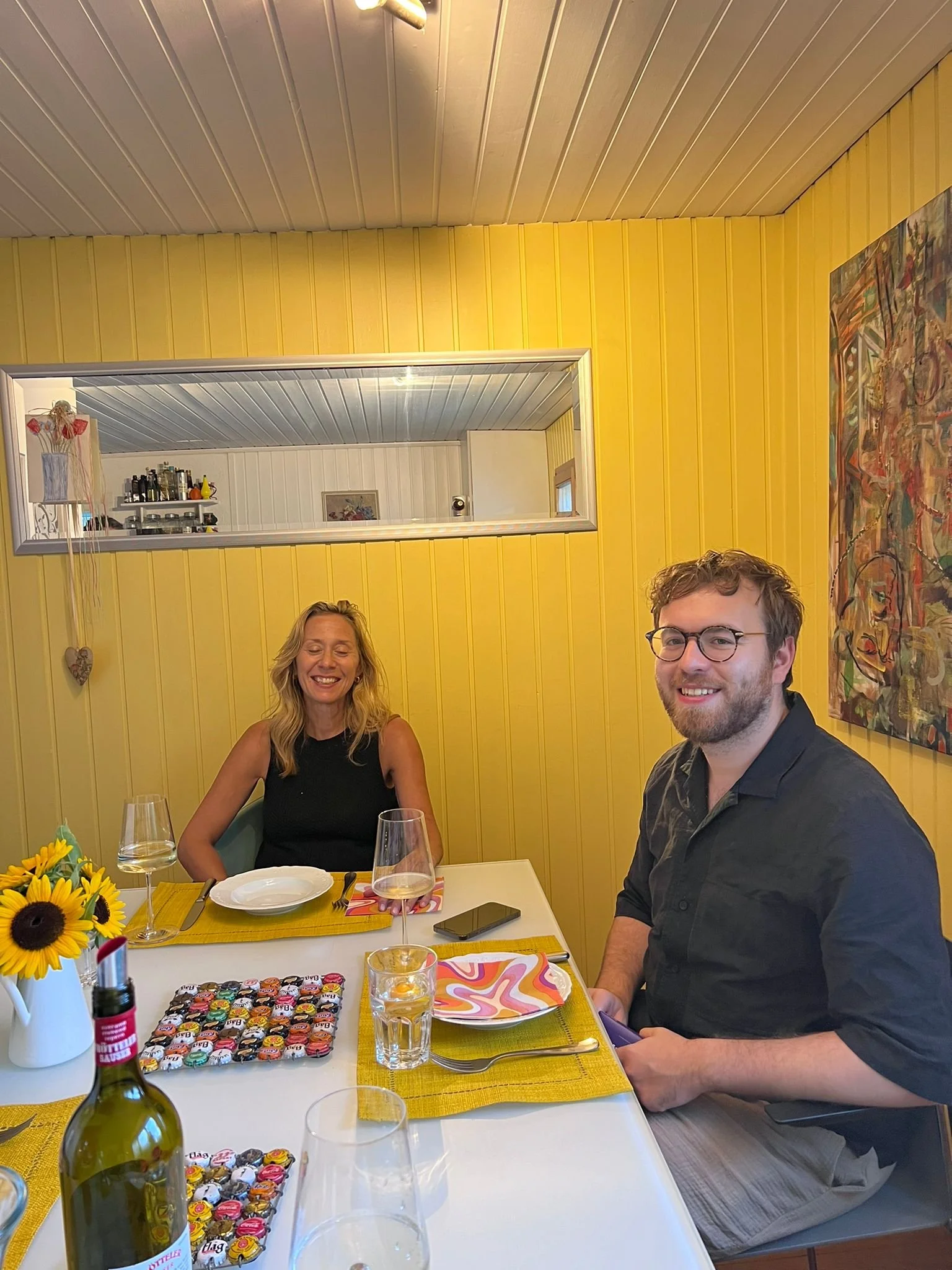 Two people sitting at a dining table, smiling, with plates, glasses, and a bottle of wine, in a room with yellow walls and colorful artwork.
