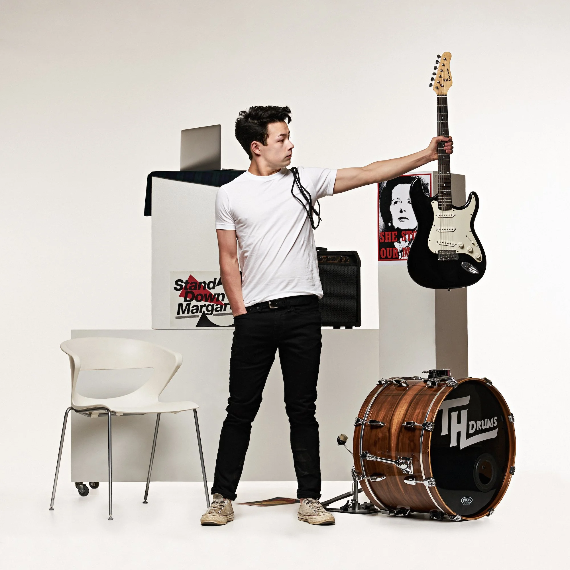 Young man standing in a music studio holding a black and white electric guitar. There is a drum set, a chair, and posters on the wall in the background.