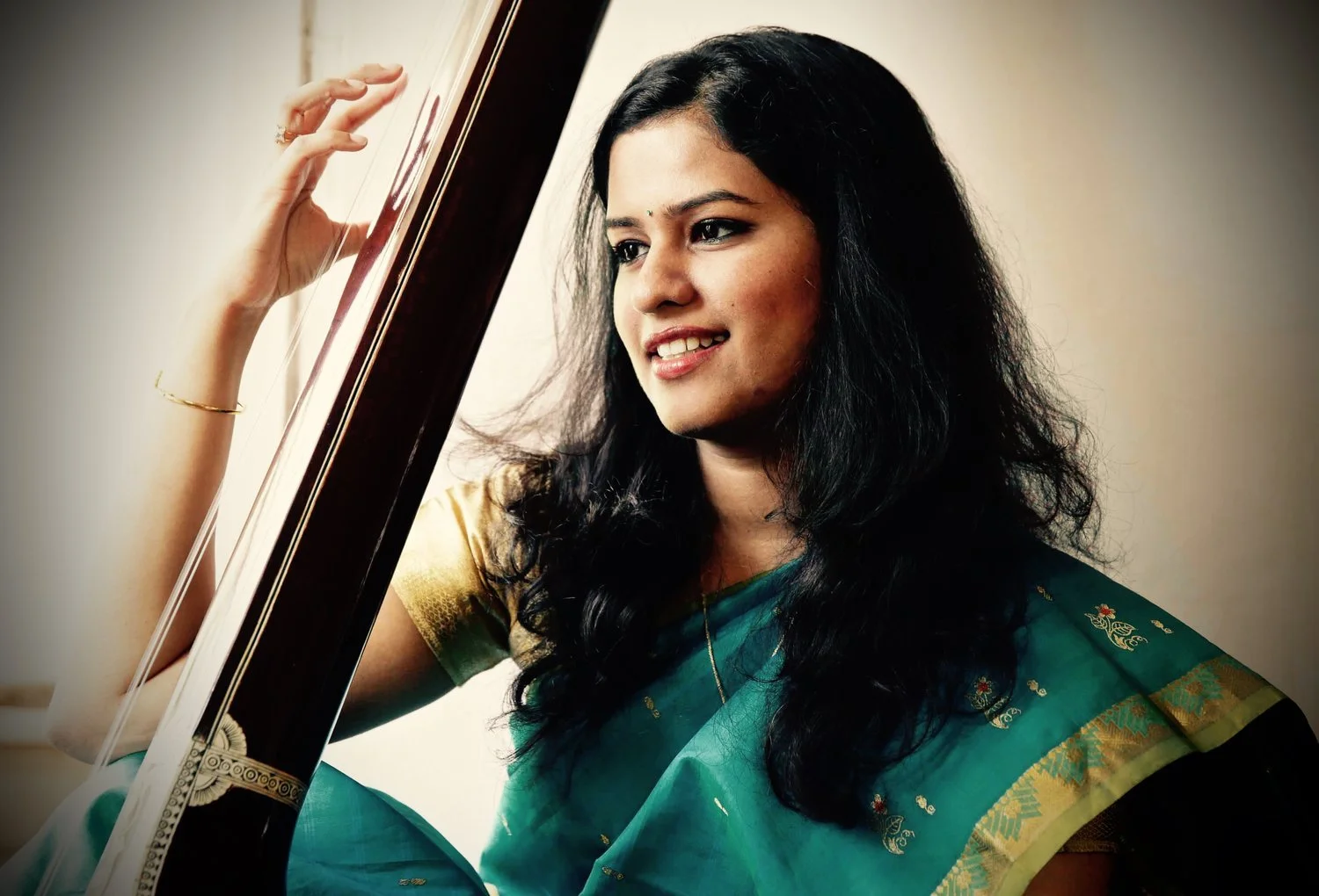 A woman with long black curly hair wearing a teal sari with gold embroidery, smiling and touching a framed mirror.