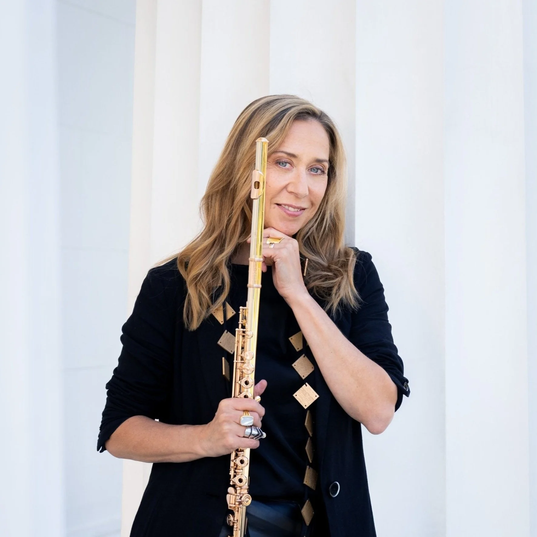 A woman with wavy blonde hair holding a gold-colored flute, standing in front of a white wall.