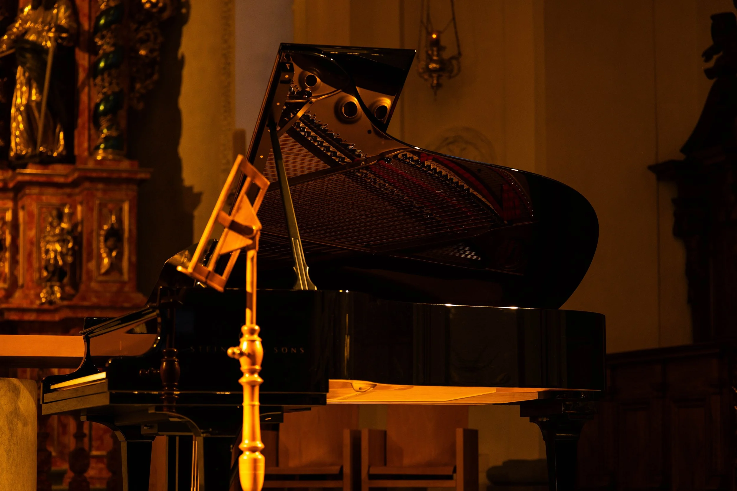 A grand piano with its lid open is in a room with warm lighting, and a brass music stand stands beside it.