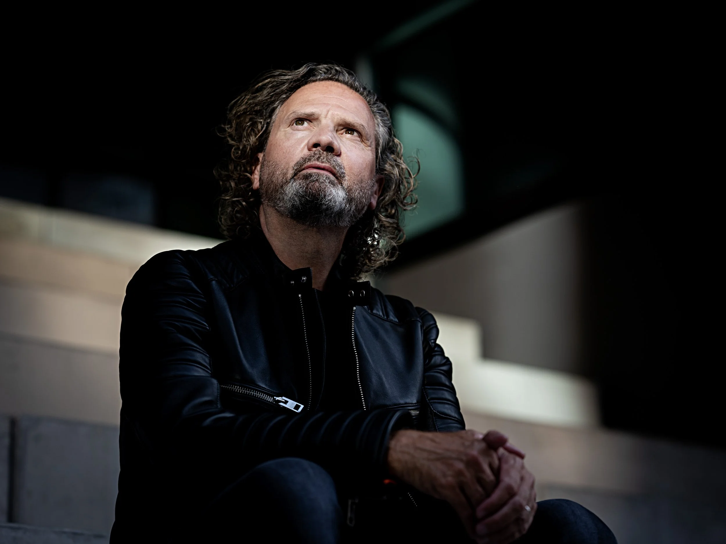 A man with curly hair and a beard, wearing a black leather jacket, sitting on a bench with a contemplative expression in a dimly lit setting.