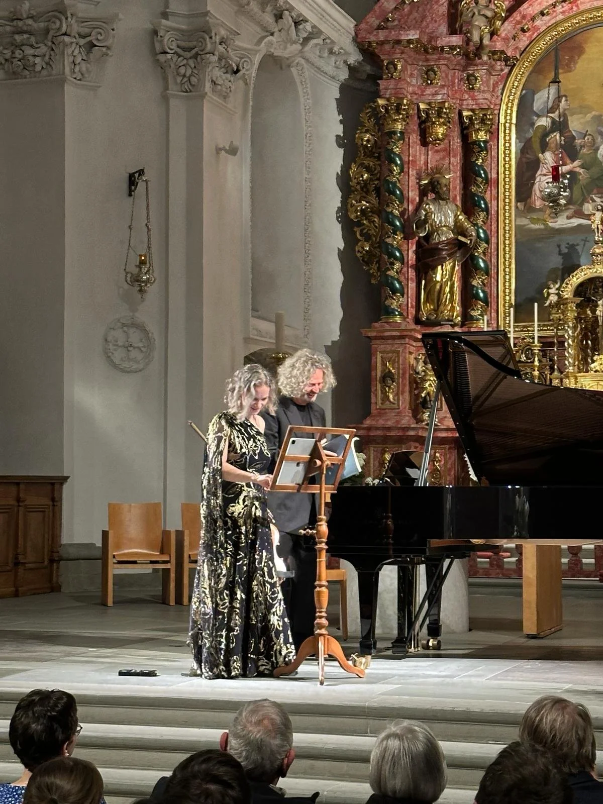 Two performers, a woman and a man, stand at a music stand in a church or concert hall, preparing for a performance by a grand piano, with an audience watching.