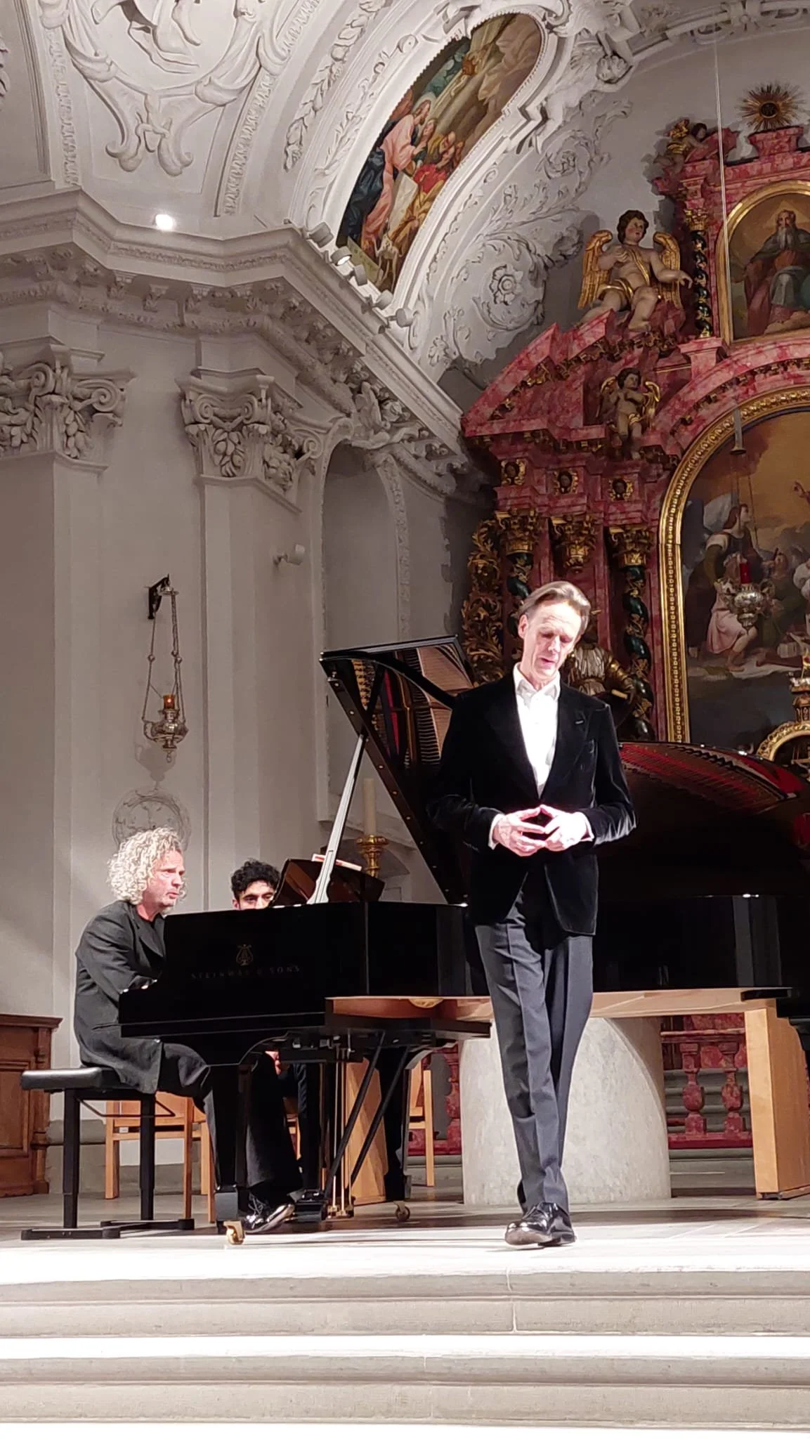A man in a black blazer stands near a grand piano while two other musicians, one with curly hair, play the piano in a church or concert hall with ornate ceiling and religious artwork.