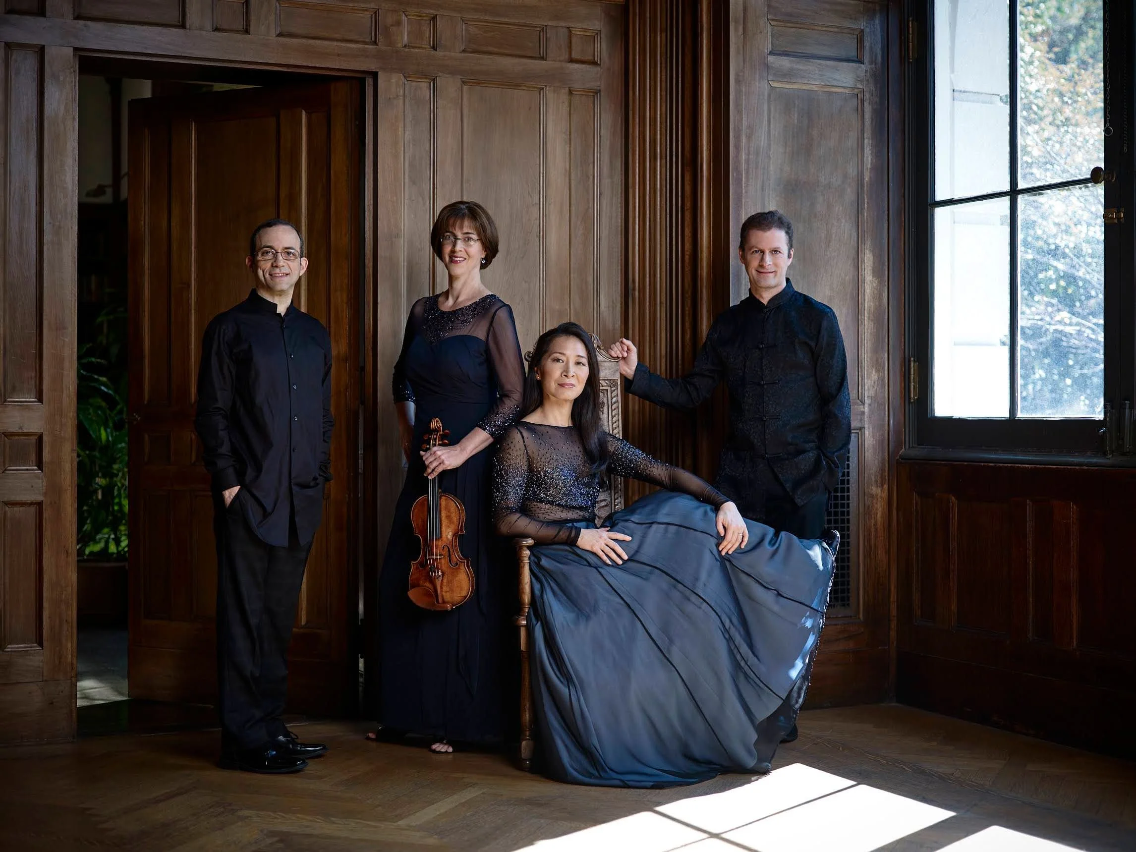 Group of four musicians in formal attire inside a wood-paneled room with large window and sunlight streaming in.