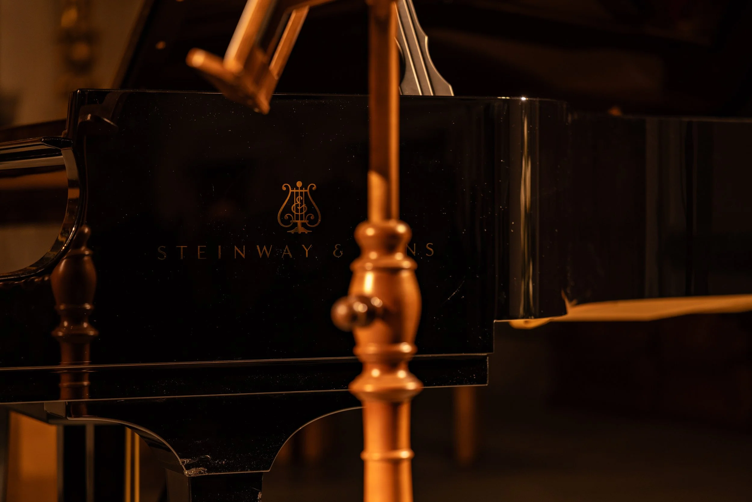 Close-up of a black Steinway & Sons grand piano with gold accents, focusing on the logo and part of the keyboard in a dimly lit room.
