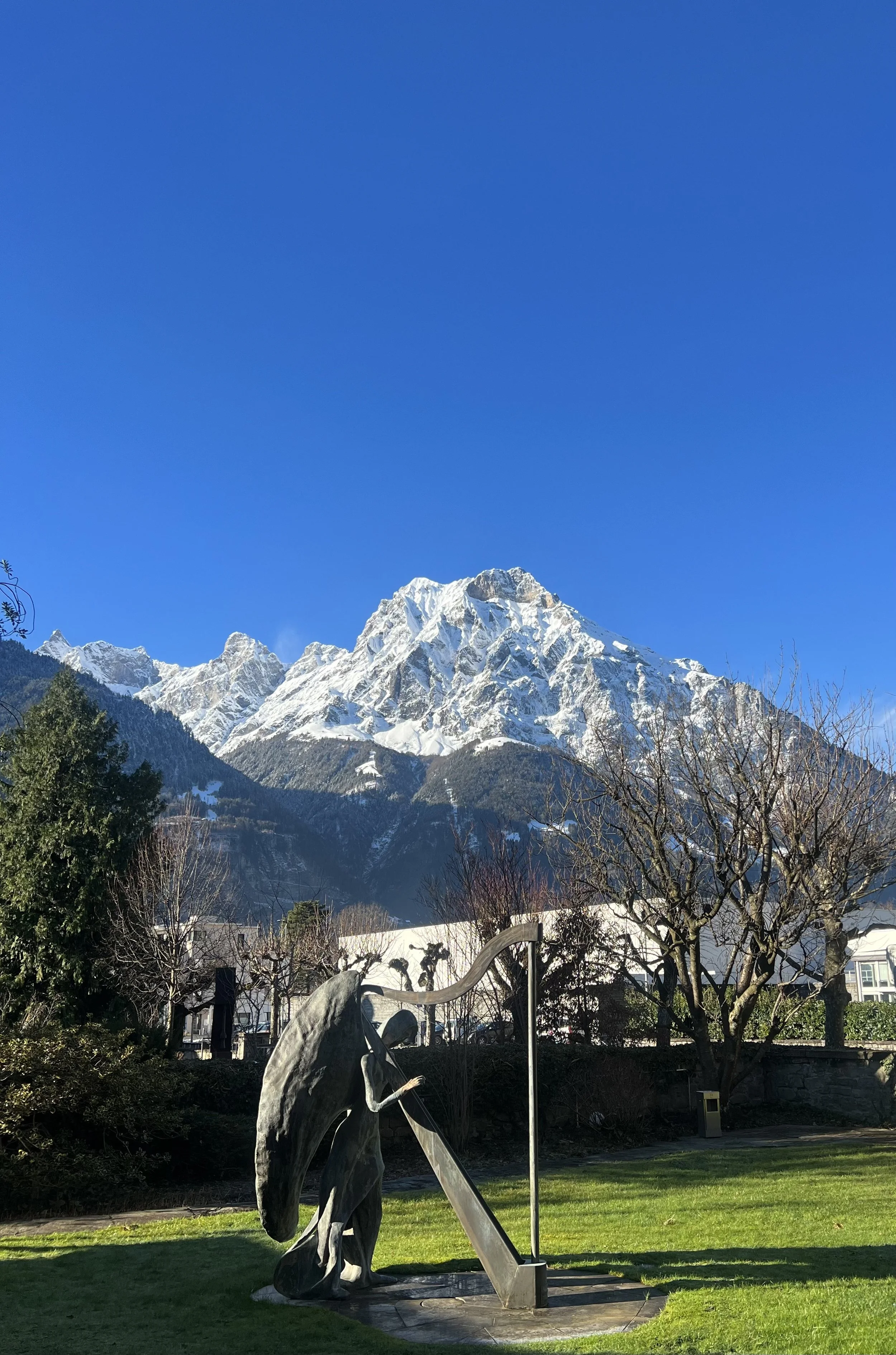 A bronze statue of a person playing a harp in a park, with snow-capped mountains and a bright blue sky in the background.