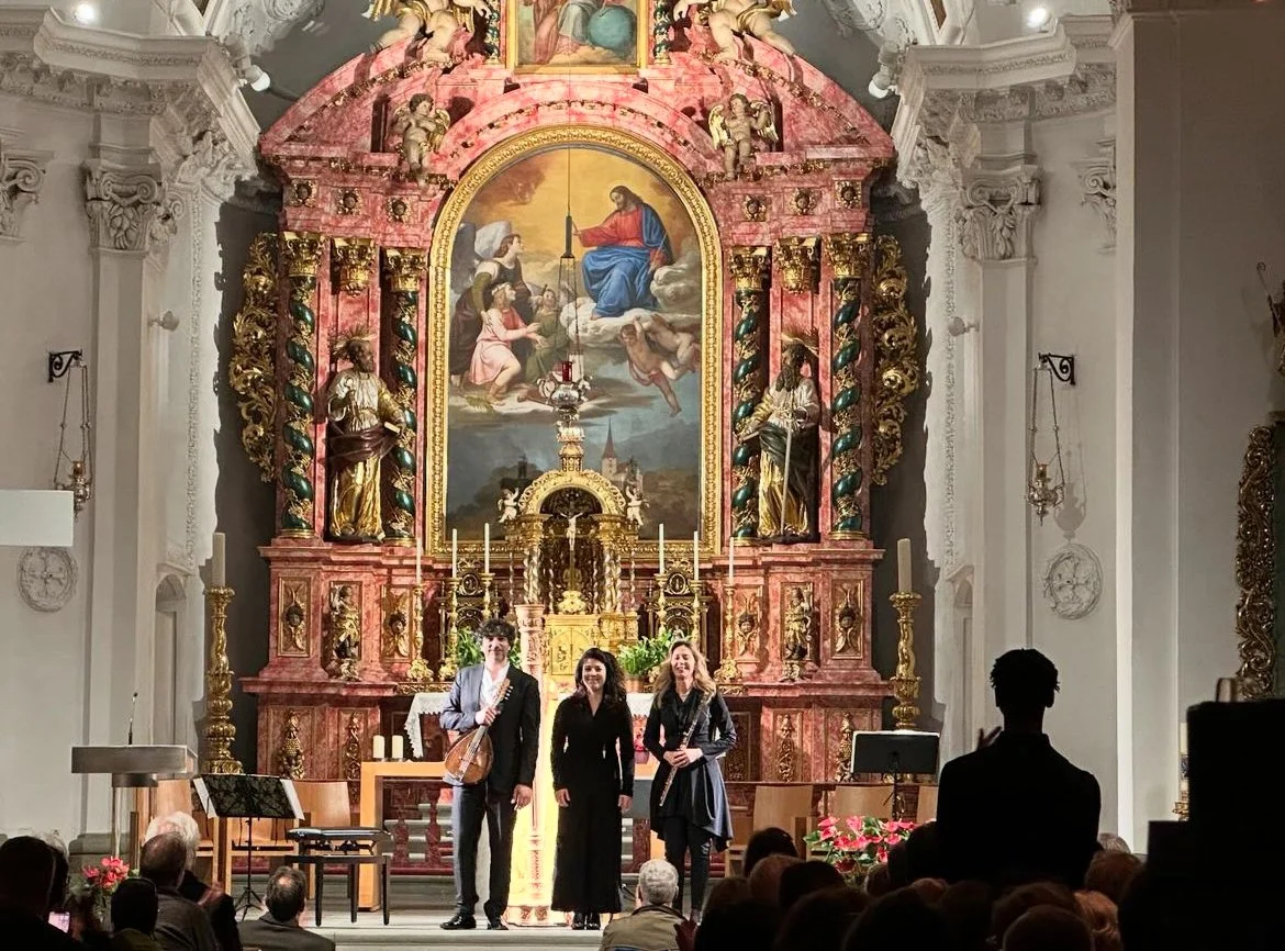 Three women stand on stage in front of an ornate church altar with religious paintings and gold accents, greeting an audience.