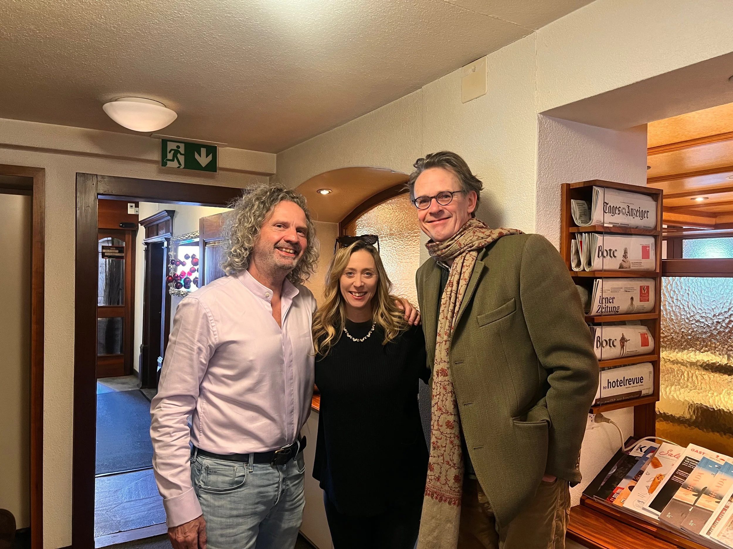 Three smiling people standing together inside a building, with a man with curly gray hair and beard on the left, a woman with long blonde hair in the middle, and a man with glasses and gray hair on the right, posing for a picture.