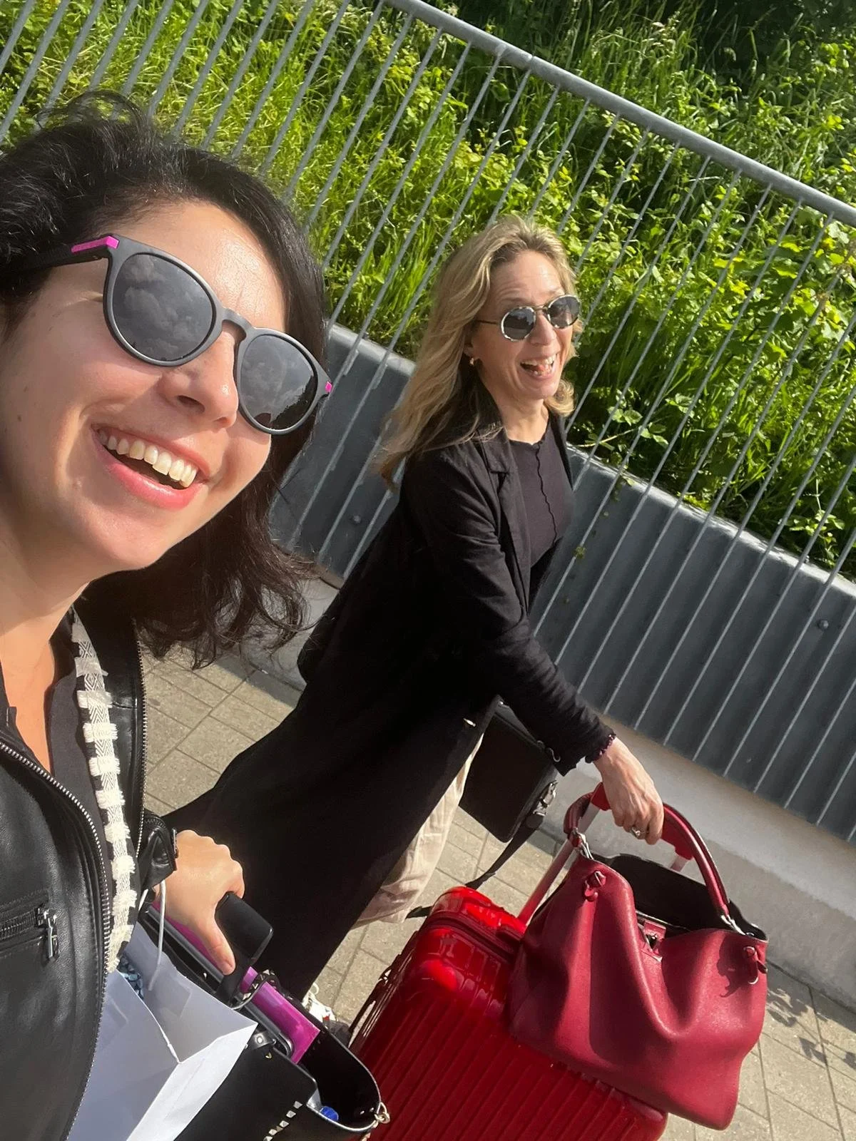 Two smiling women wearing sunglasses and black clothing, walking outdoors with luggage, on a paved path next to a metal fence and green foliage.