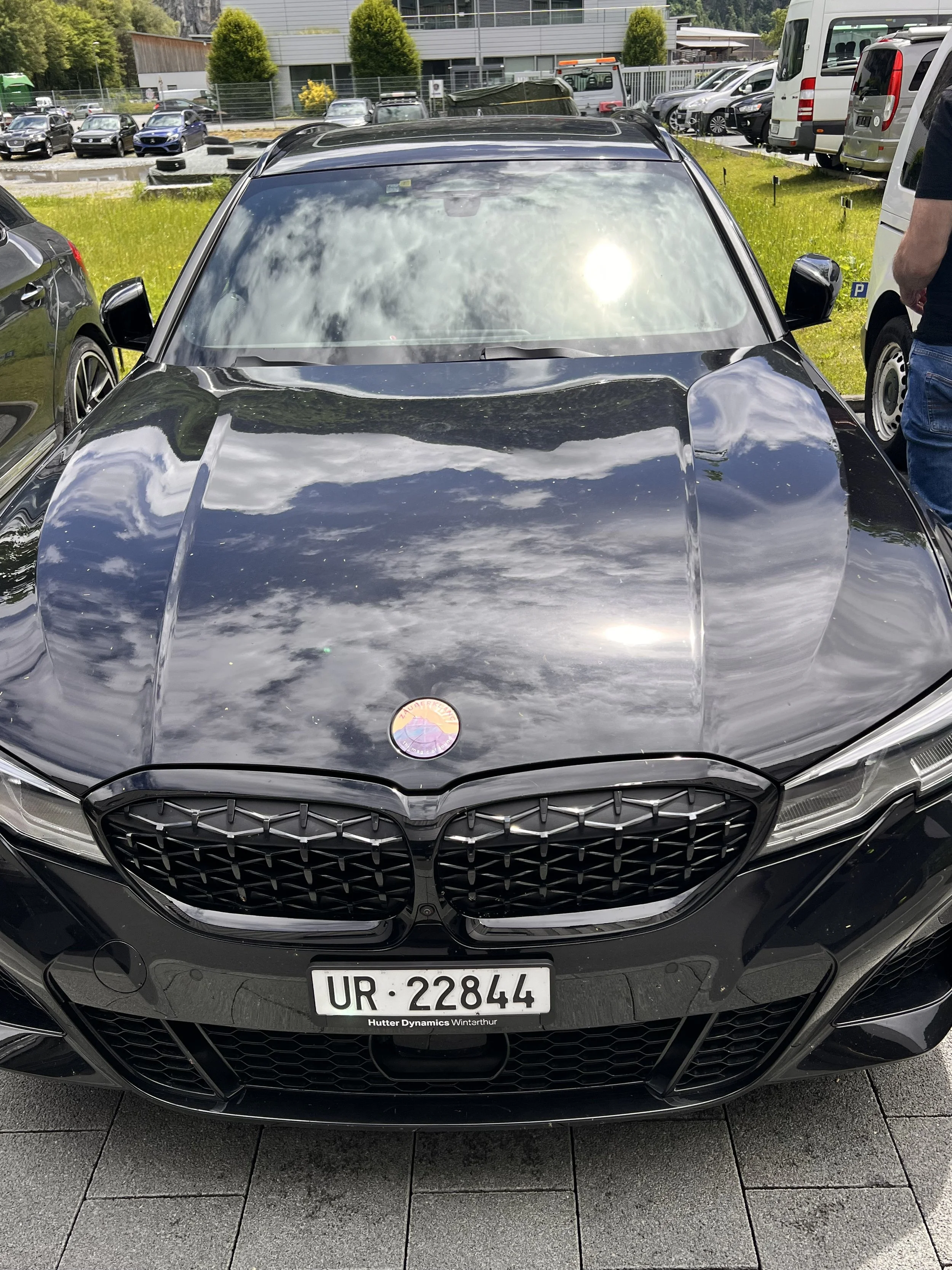 Front view of a black car, reflecting the cloudy sky, parked in a lot with other vehicles and yellow-green grass in the background.