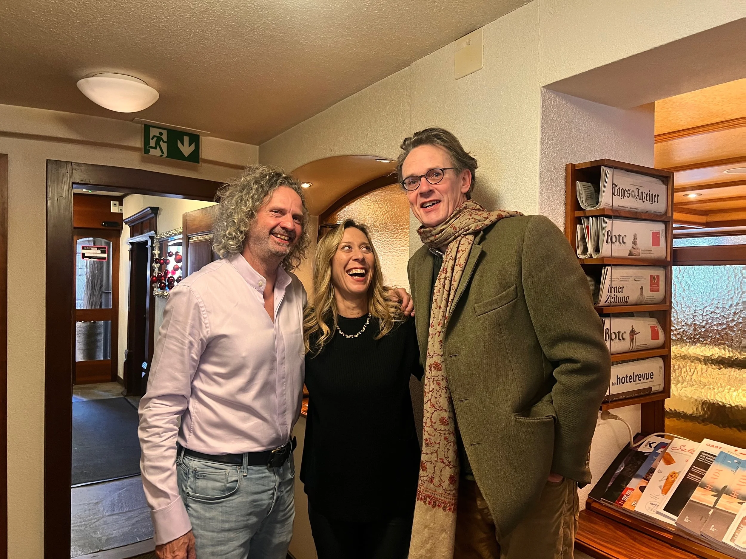 Three people are smiling and posing together inside a building. A man in a white shirt and jeans, a woman in a black top, and a man in a green blazer with glasses and a scarf. There are newspapers and magazines on a table and a sign pointing down on