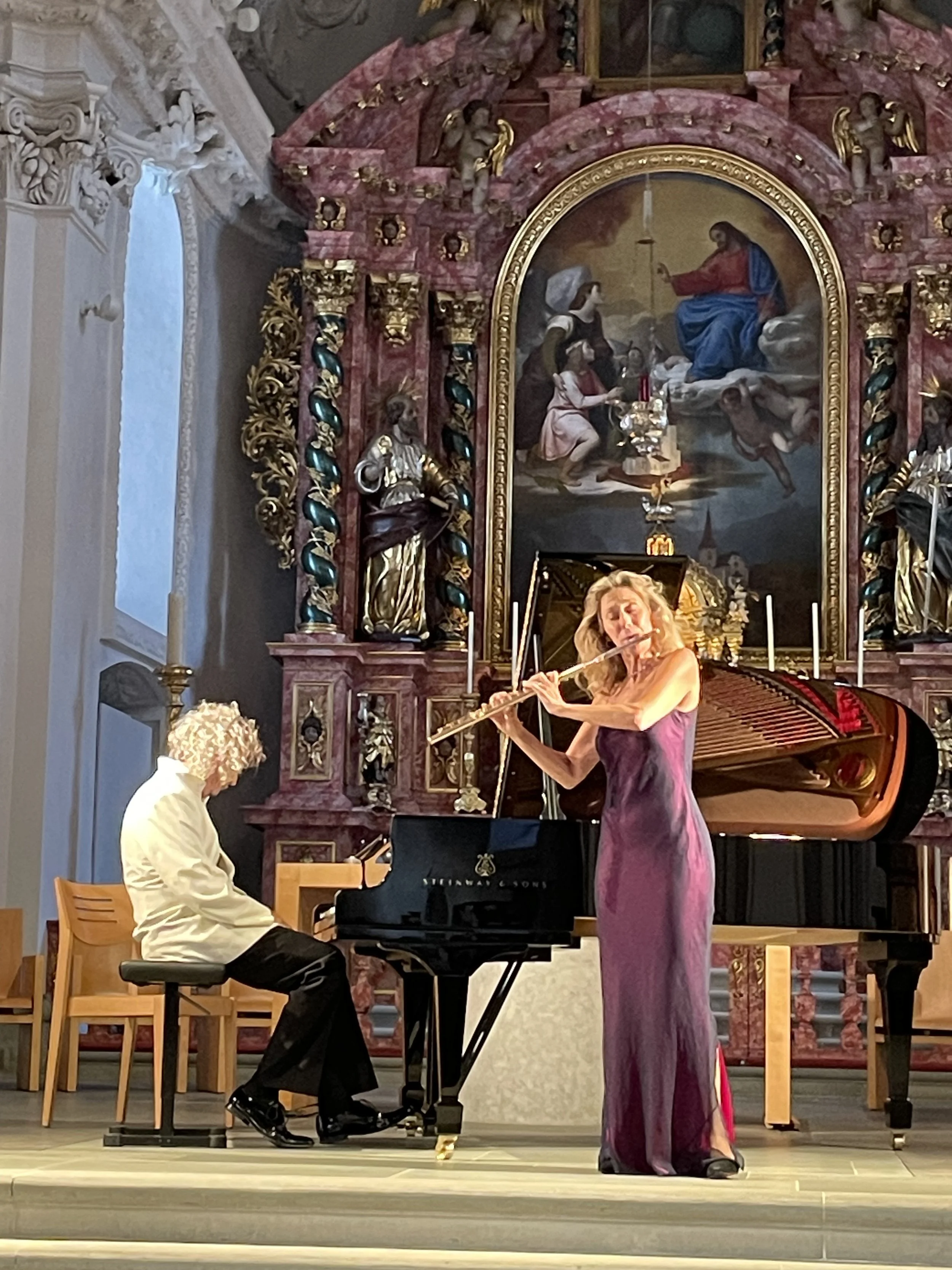Musicians performing on stage inside a church, with a woman playing the flute and a man playing the piano, in front of a large religious painting and ornate altar.