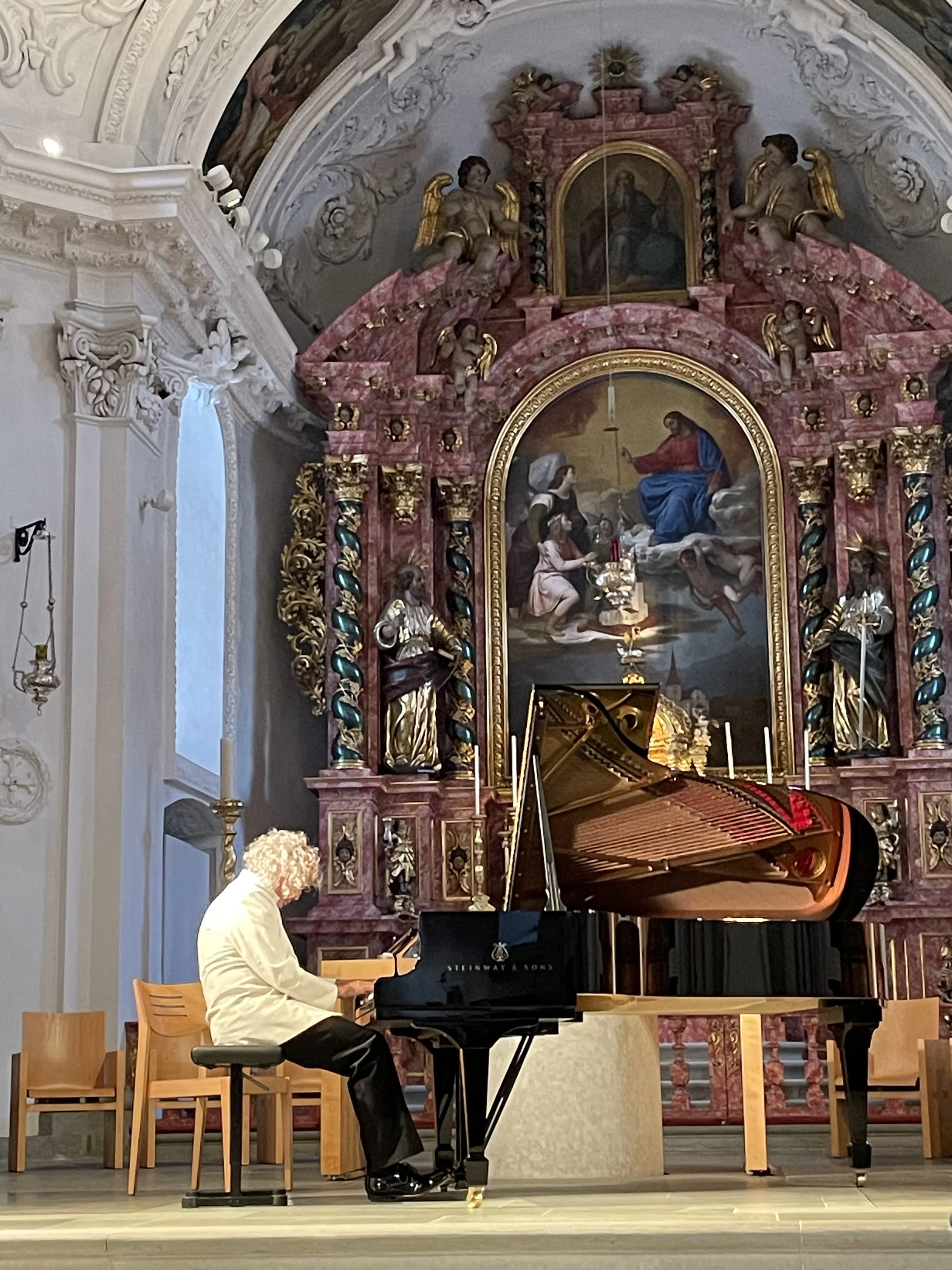 A person with curly gray hair playing a grand piano on a stage inside a church or cathedral, with an ornate altar featuring religious paintings and sculptures in the background.