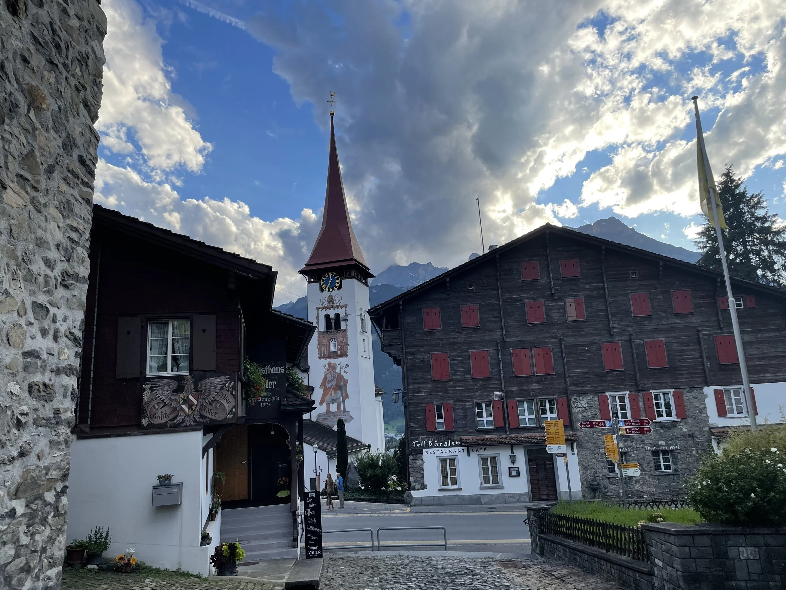 Colorful European village street scene with a church with a tall steeple, old-style buildings, signs, and mountains in the background under a partly cloudy sky.
