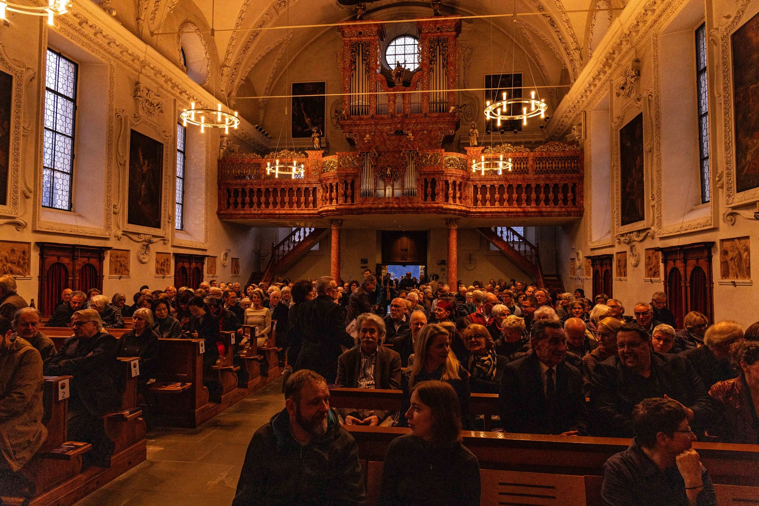 A large group of people seated in a church or cathedral, waiting for an event to start. The interior features ornate woodwork, a pipe organ at the back, and high arched ceilings.
