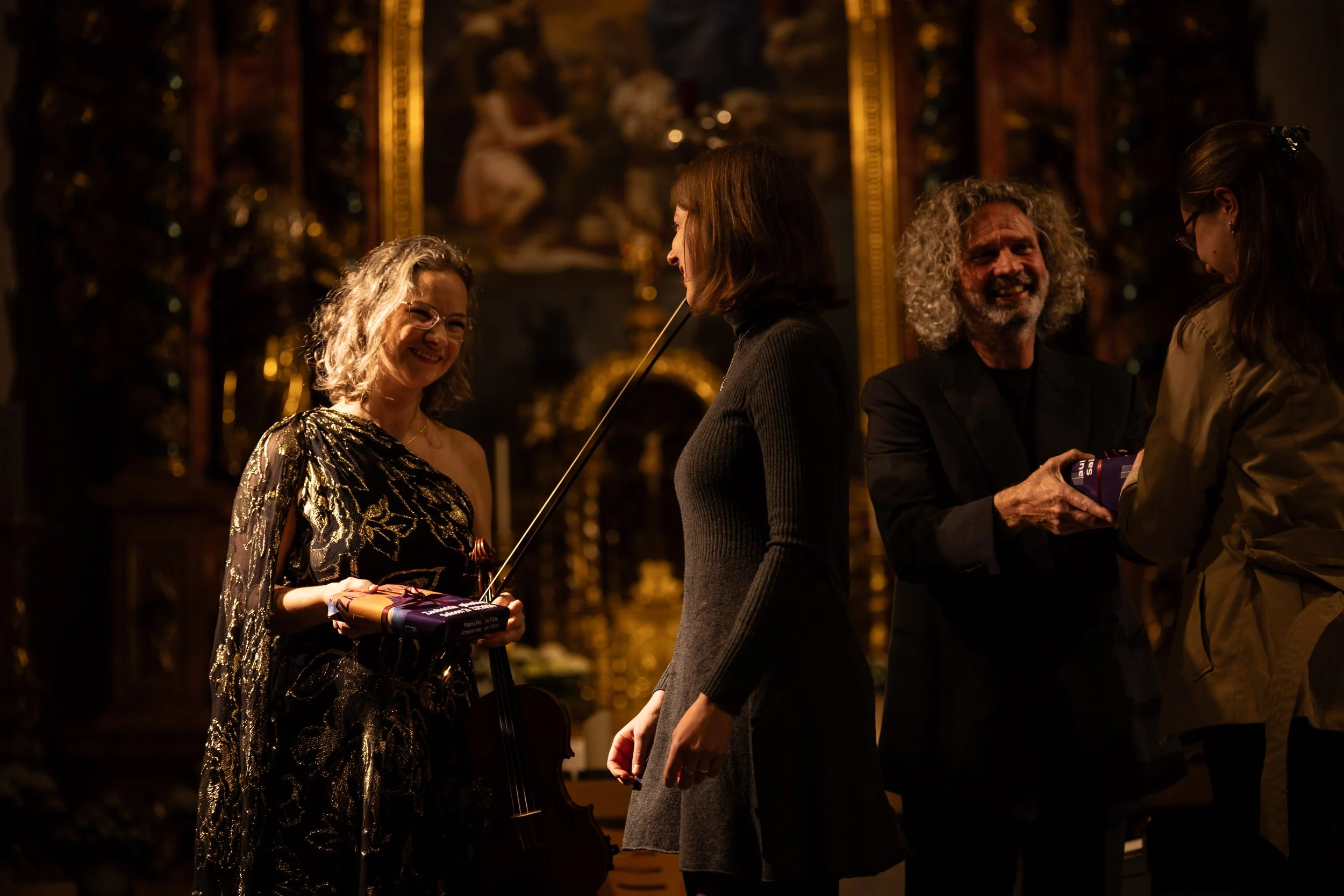 Four people are inside a dimly lit, ornate room, engaging in conversation and exchanging items. The woman on the left is smiling, holding a small wrapped gift and a music stick, with a violin at her side. The man on the right has curly gray hair and
